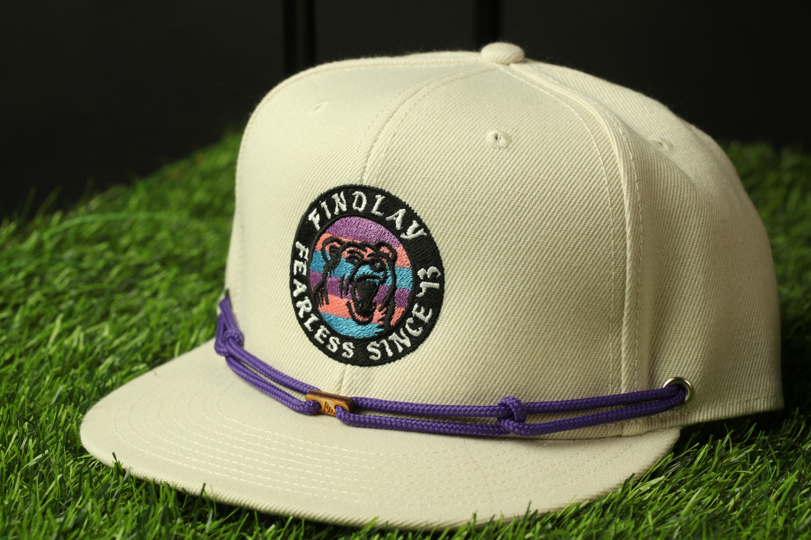 A beige baseball cap with a purple and black embroidered logo, sitting on a grassy surface.