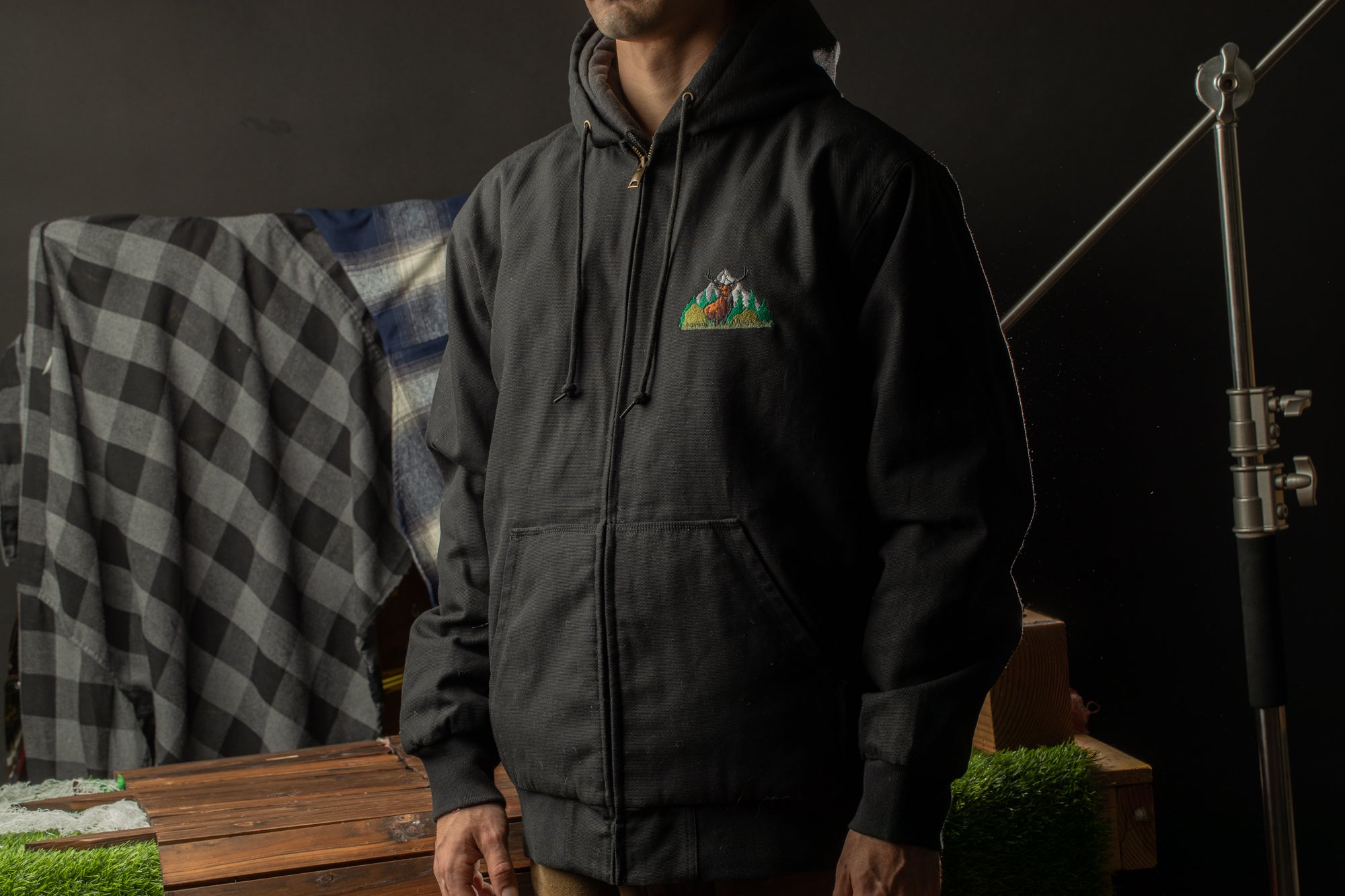 A person wearing a black hooded jacket with a mountain-like logo stands in a dark room with a checkered blanket and a wooden table in the background.