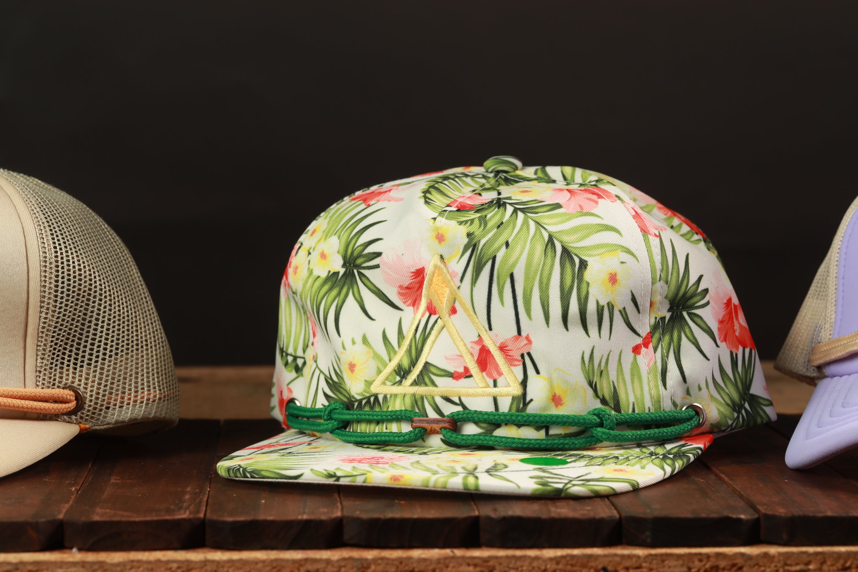 A floral print baseball cap with a green band is displayed on a wooden surface, with other hats visible in the background.