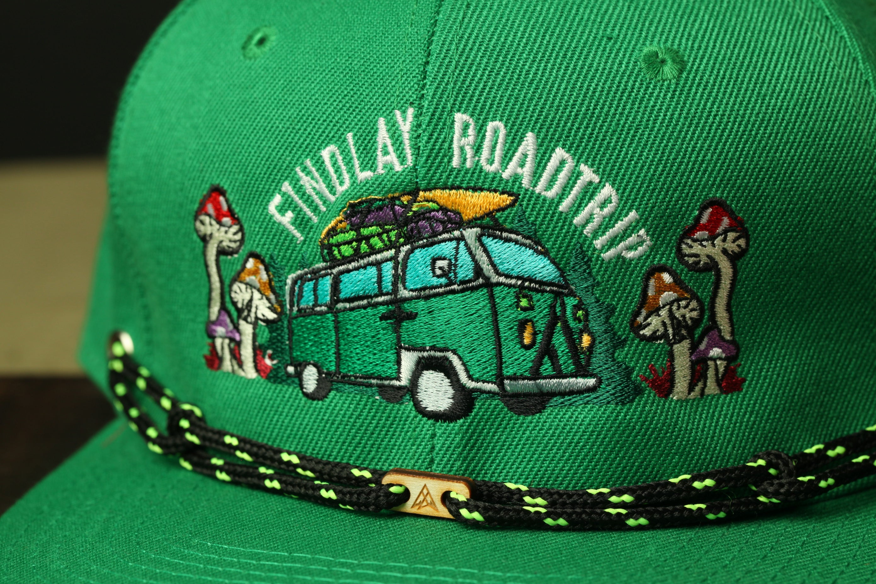 A close-up view of a green baseball cap with an embroidered design featuring a van, mushrooms, and the text "Findlay Road Trip".