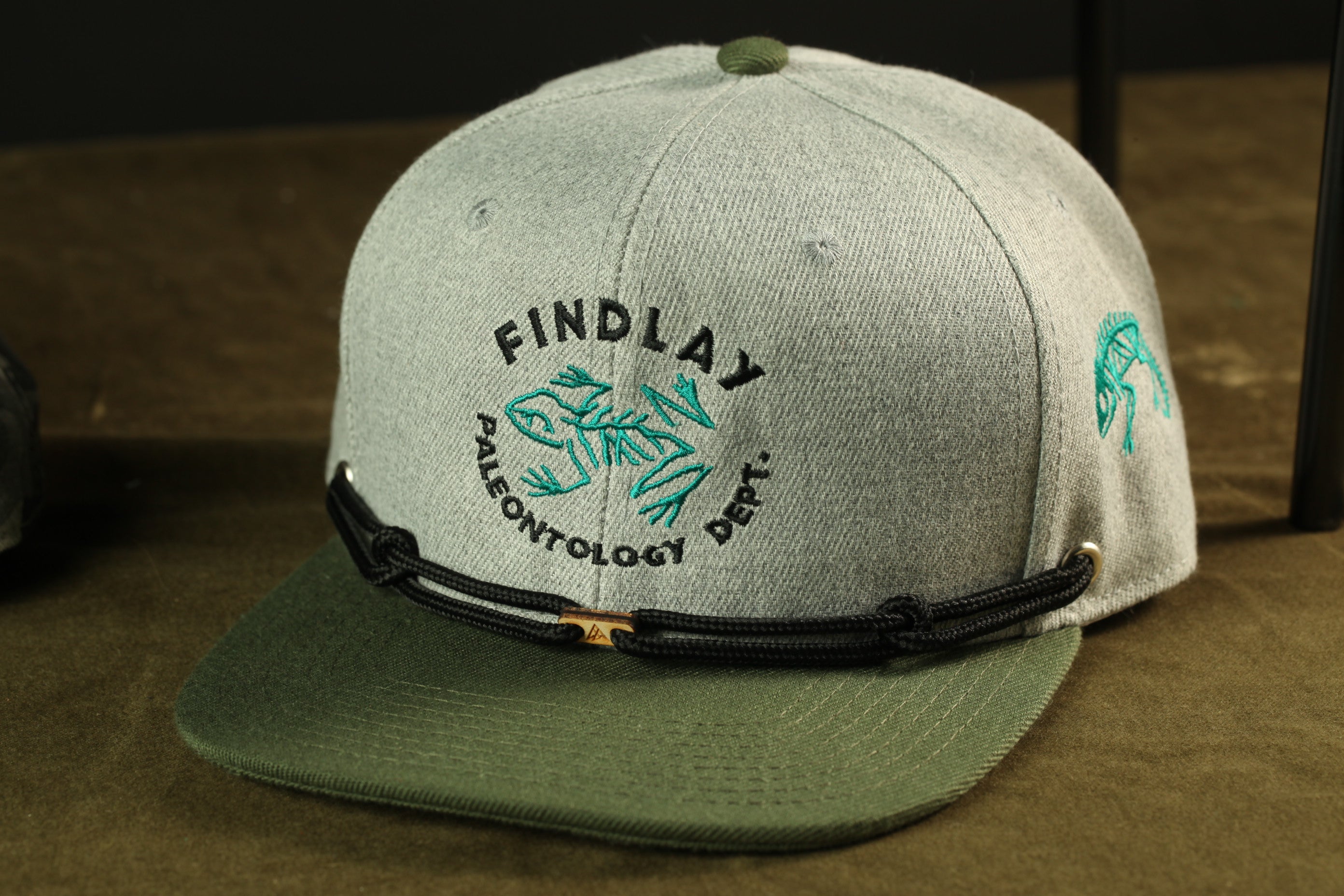 A grey baseball cap with a green and black design, featuring the text "FINDLAY PALEONTOLOGY DEPT."