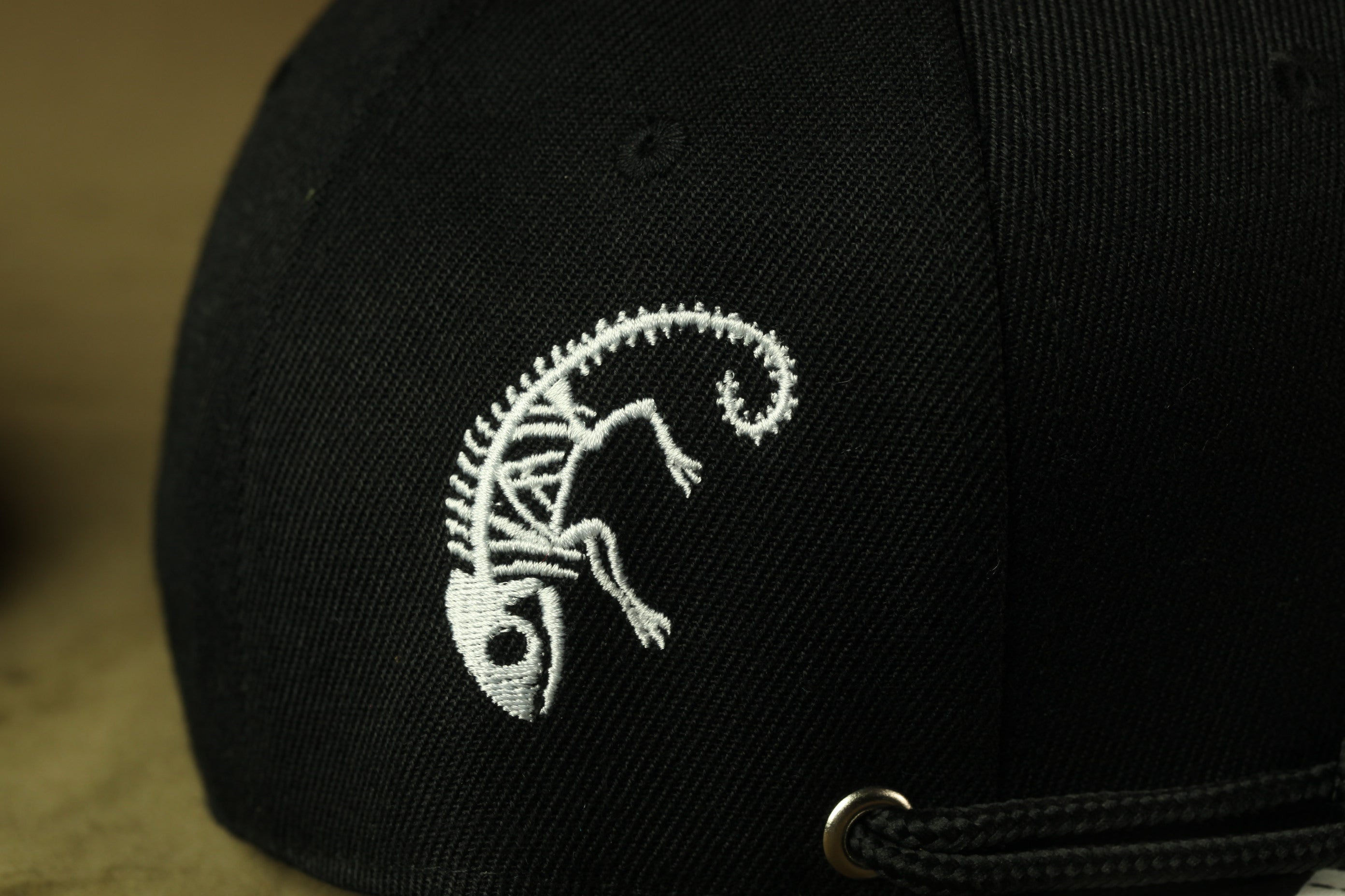 A close-up view of a black baseball cap with a white embroidered design of a skeleton or chameleon on the front.