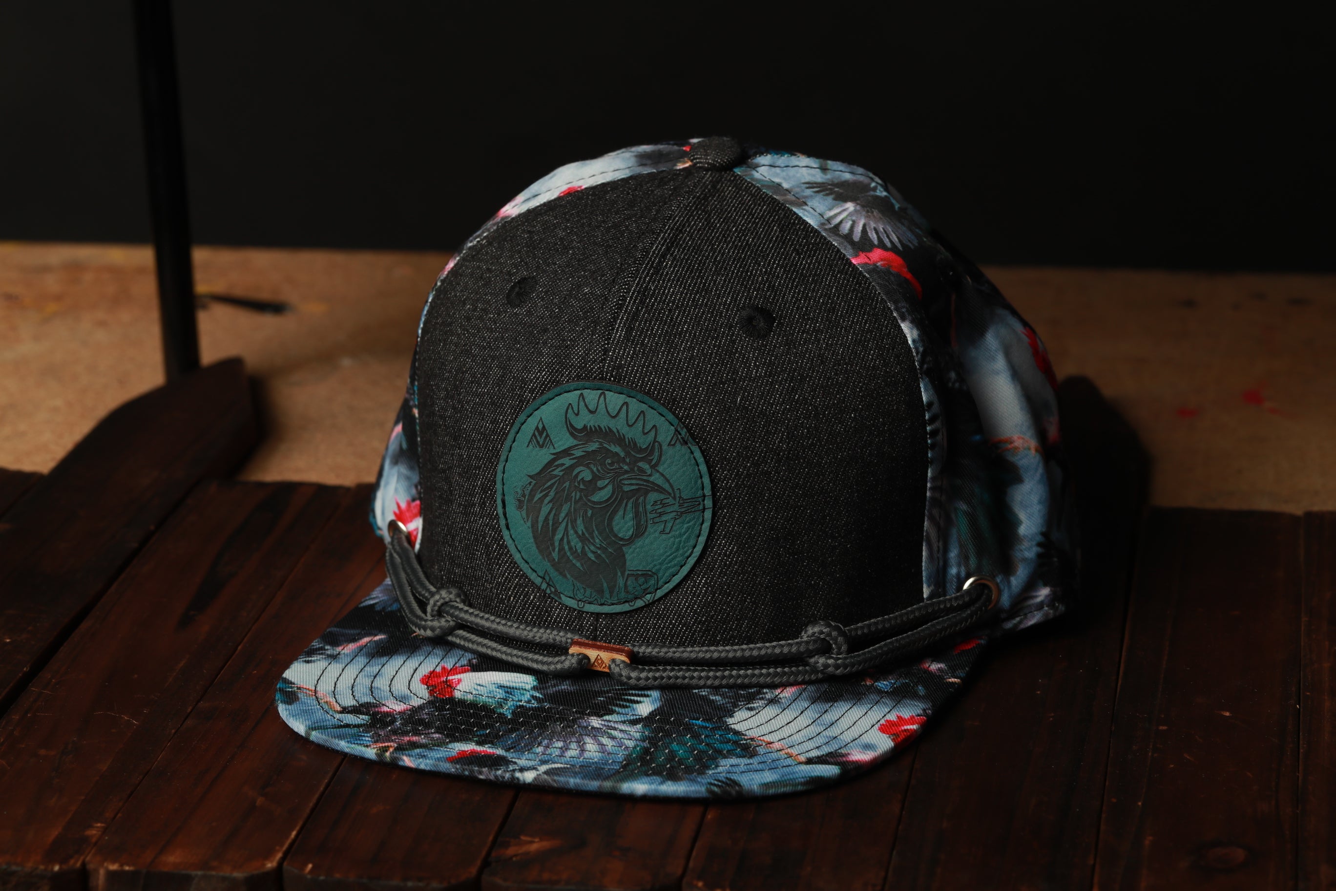 A black baseball cap with a colorful floral pattern and a circular patch on the front, placed on a wooden surface.