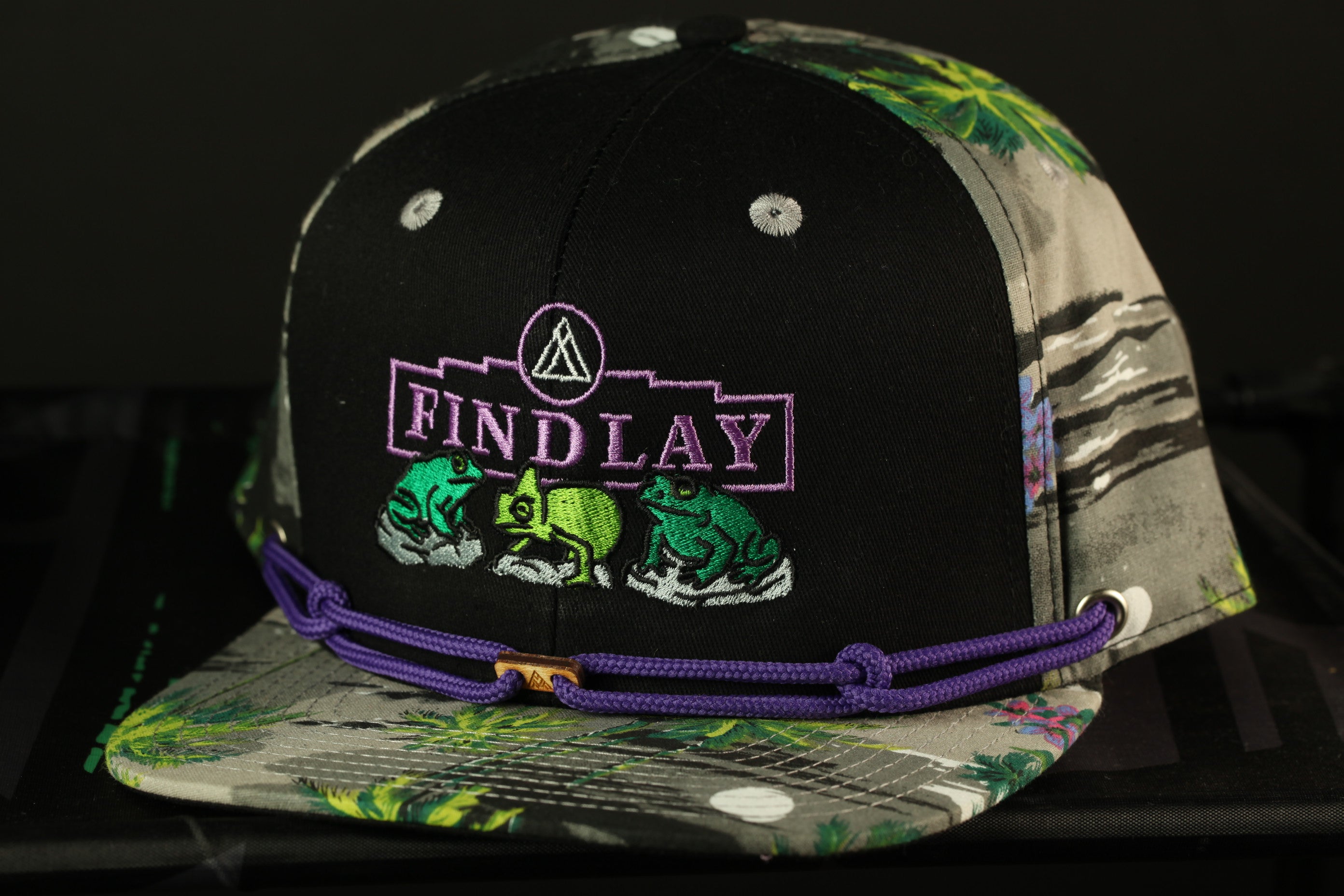 A black baseball cap with a floral pattern and purple braided strap, featuring a logo and text on the front.