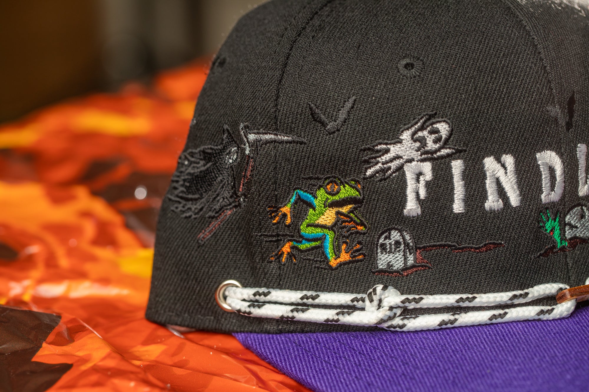 A close-up view of a black baseball cap with various embroidered designs, including a frog, a ghost, and a skull, against a background of orange and brown fabric.