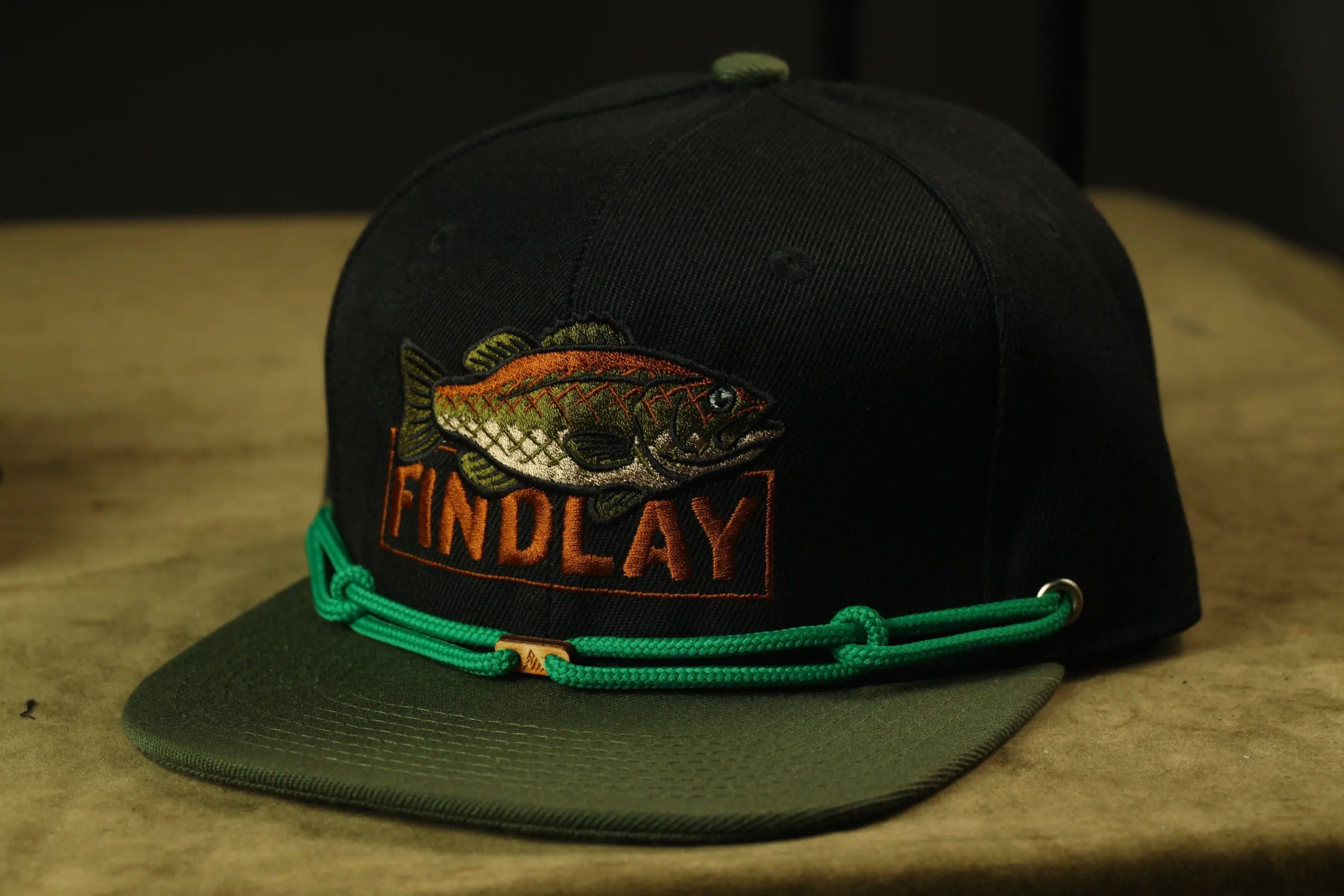 A black baseball cap with a fish embroidered on it, and a green string around the brim.