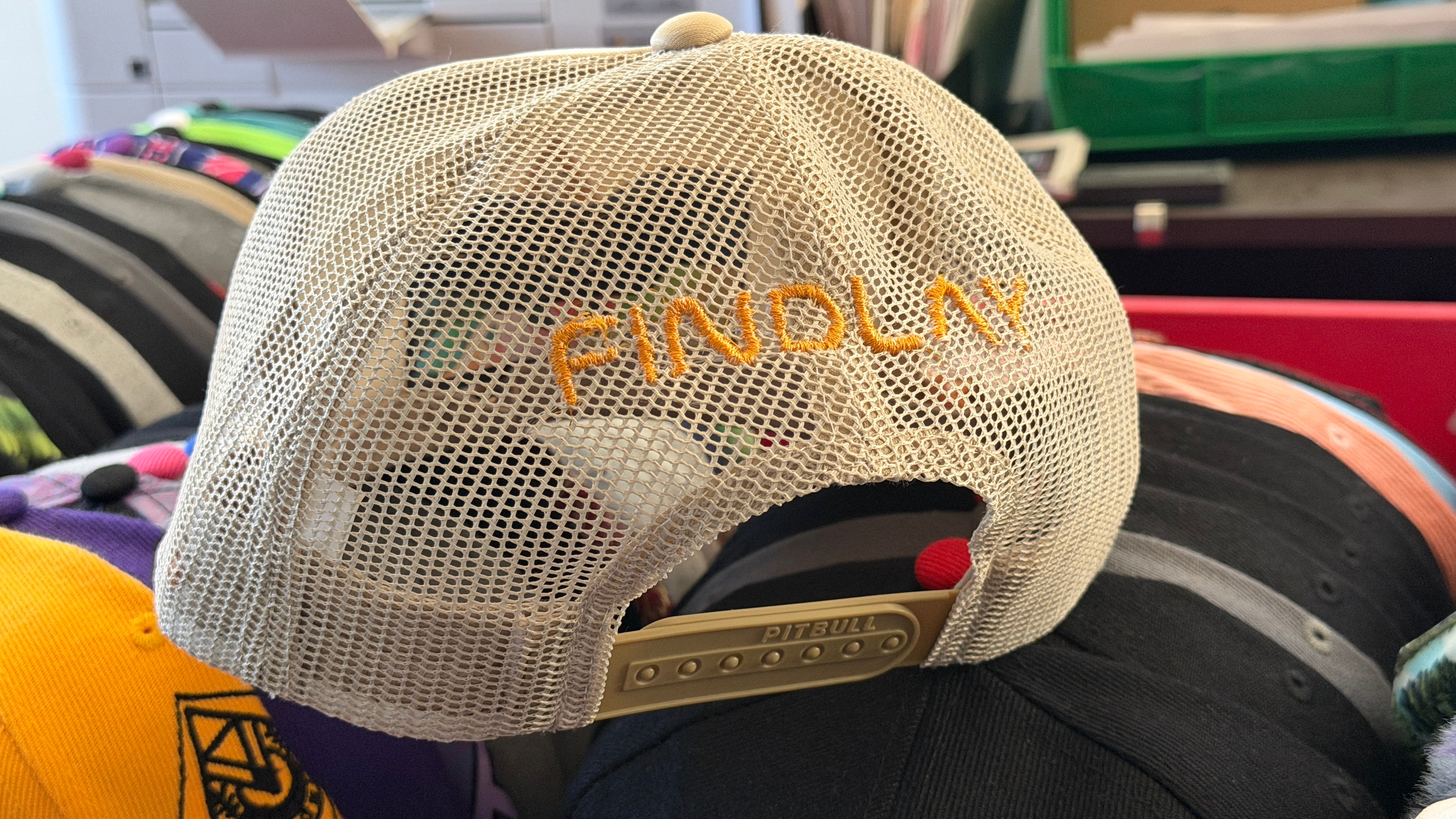A close-up view of a beige baseball cap with the word "FINDlay" embroidered on it, displayed among other baseball caps.