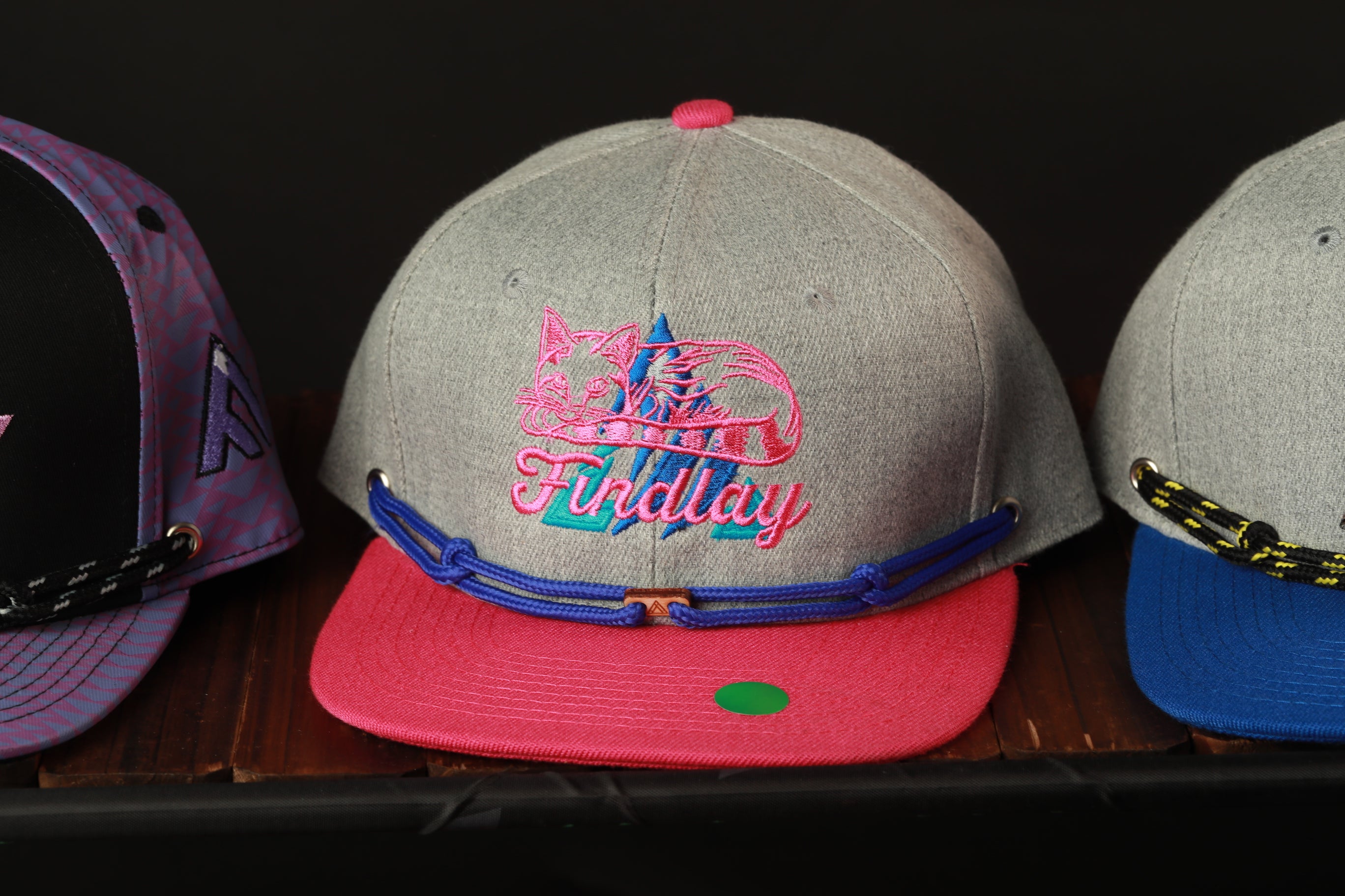 Three colorful baseball caps are displayed on a shelf, with the gray cap featuring a pink cat logo and the other two caps in different colors.