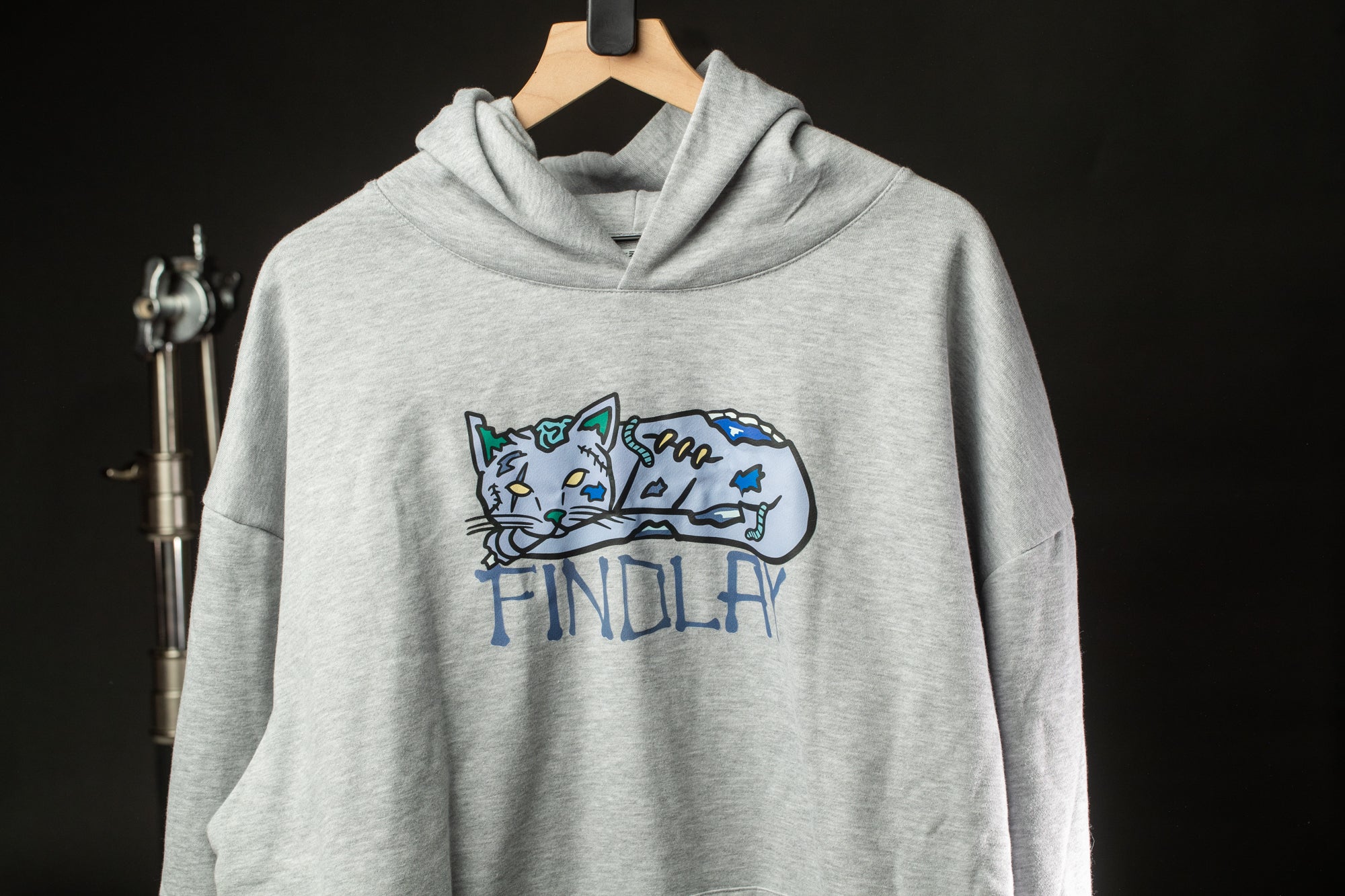 A grey hoodie with a cat graphic and the word "FINDLAY" printed on it, hanging on a wooden hanger against a dark background.