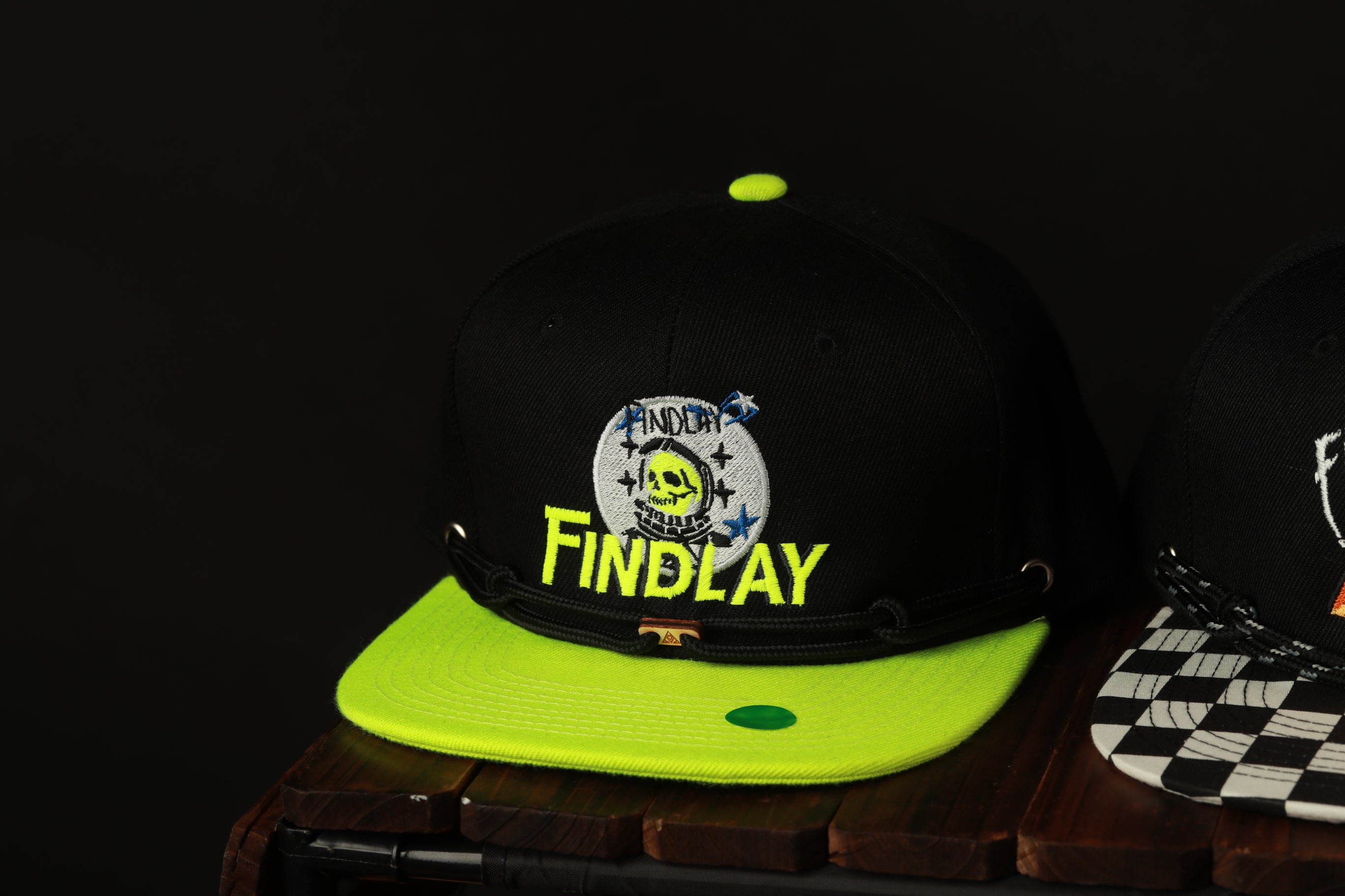 A black baseball cap with a yellow brim and a black and white checkered pattern, featuring a skull and crossbones design.