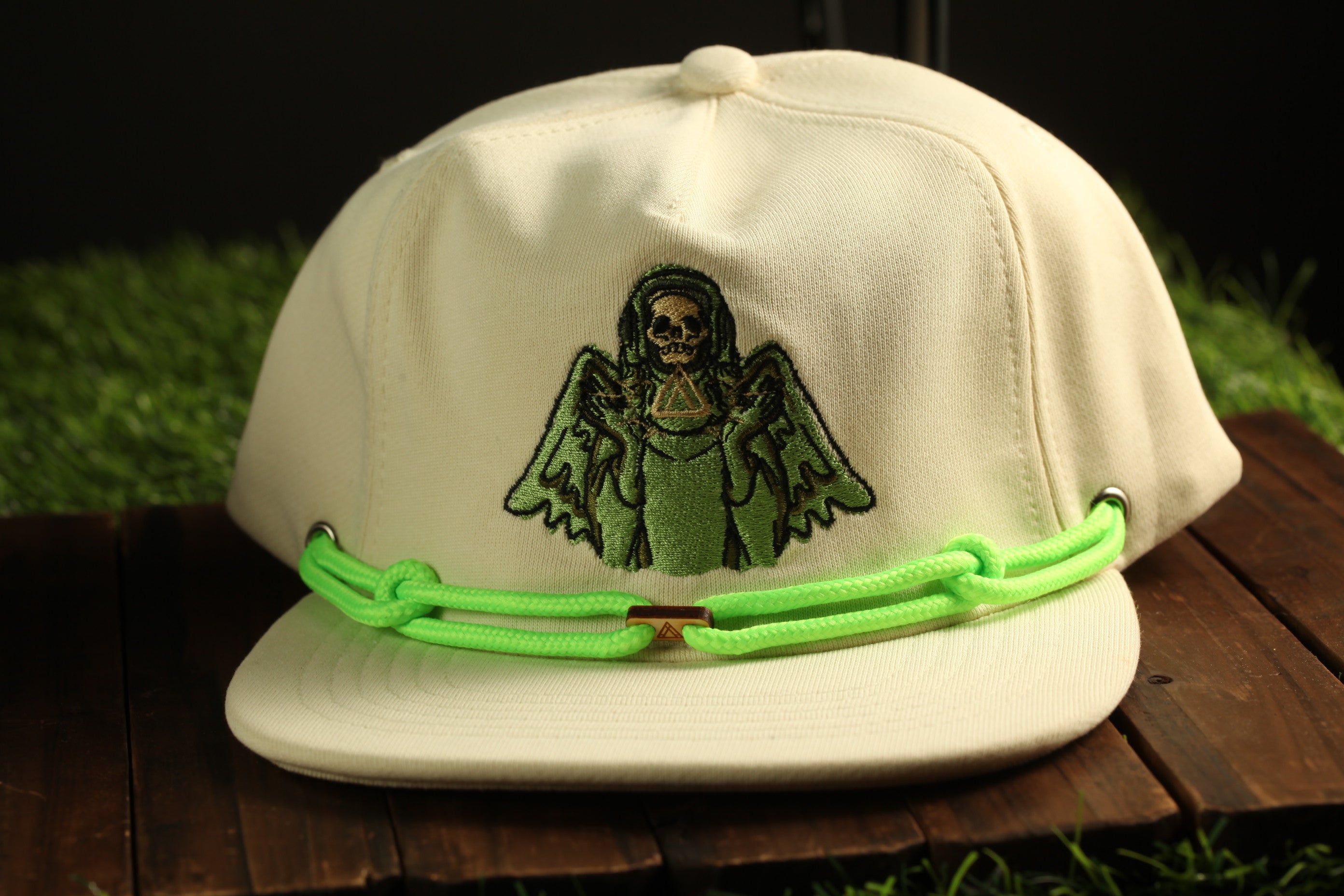 A white baseball cap with a green strap and an embroidered design of a skull and wings on the front.