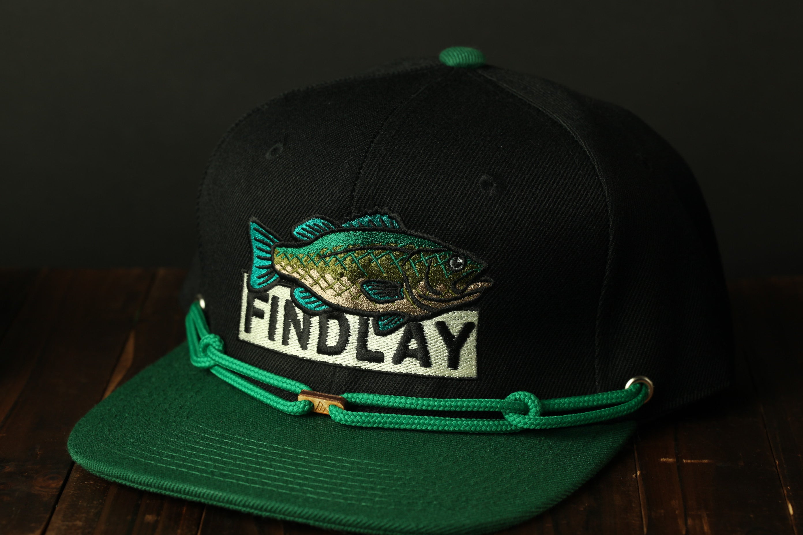 A black baseball cap with a green brim and a fish embroidered on it, sitting on a wooden surface.