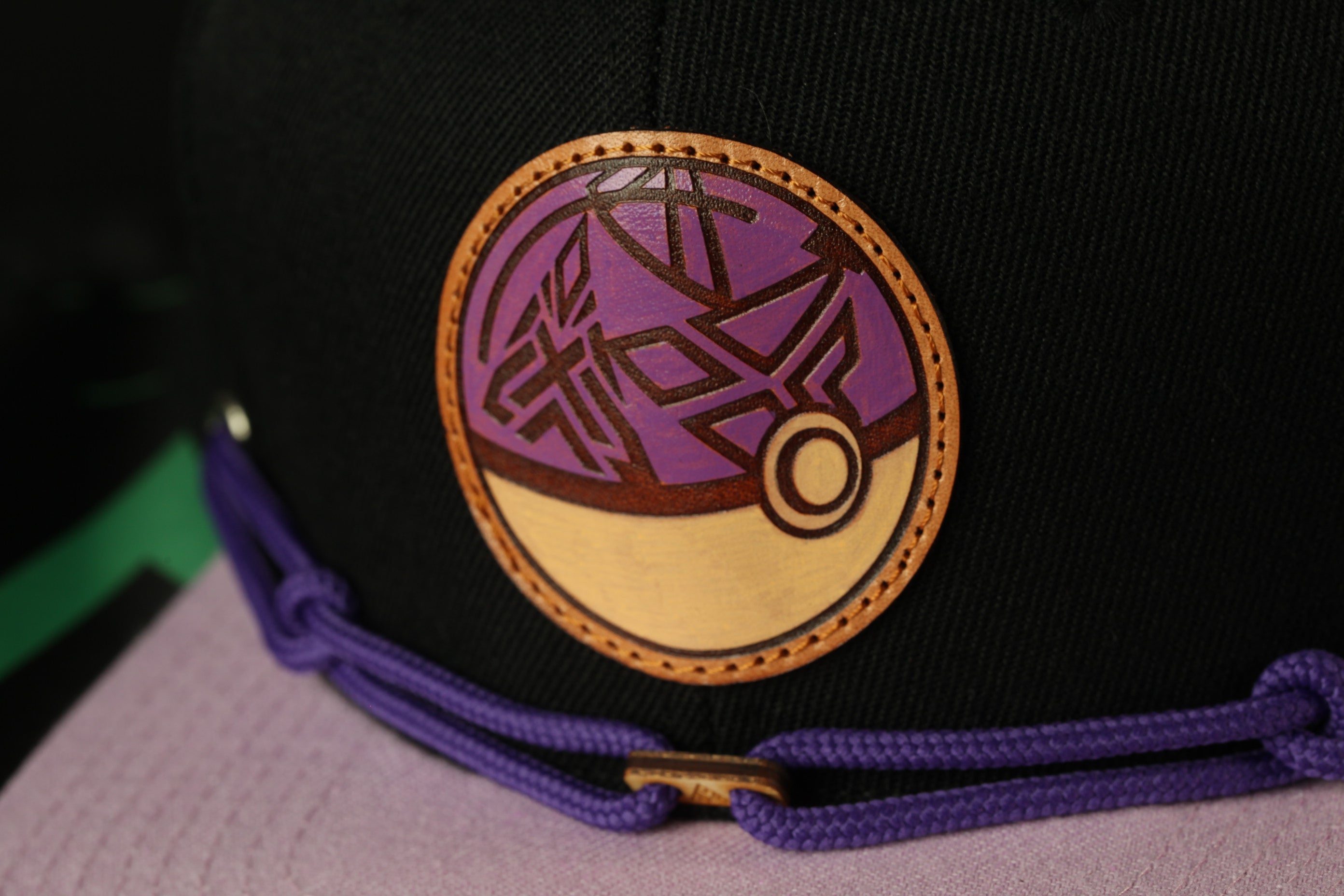 A close-up view of a black baseball cap with a purple and gold circular logo on the front.