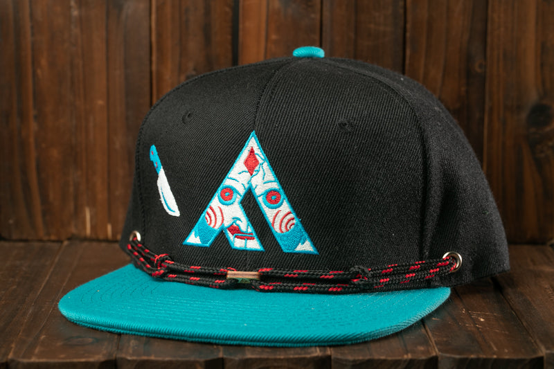 A black baseball cap with a teal brim and a blue and white graphic design on the front, sitting on a wooden surface.