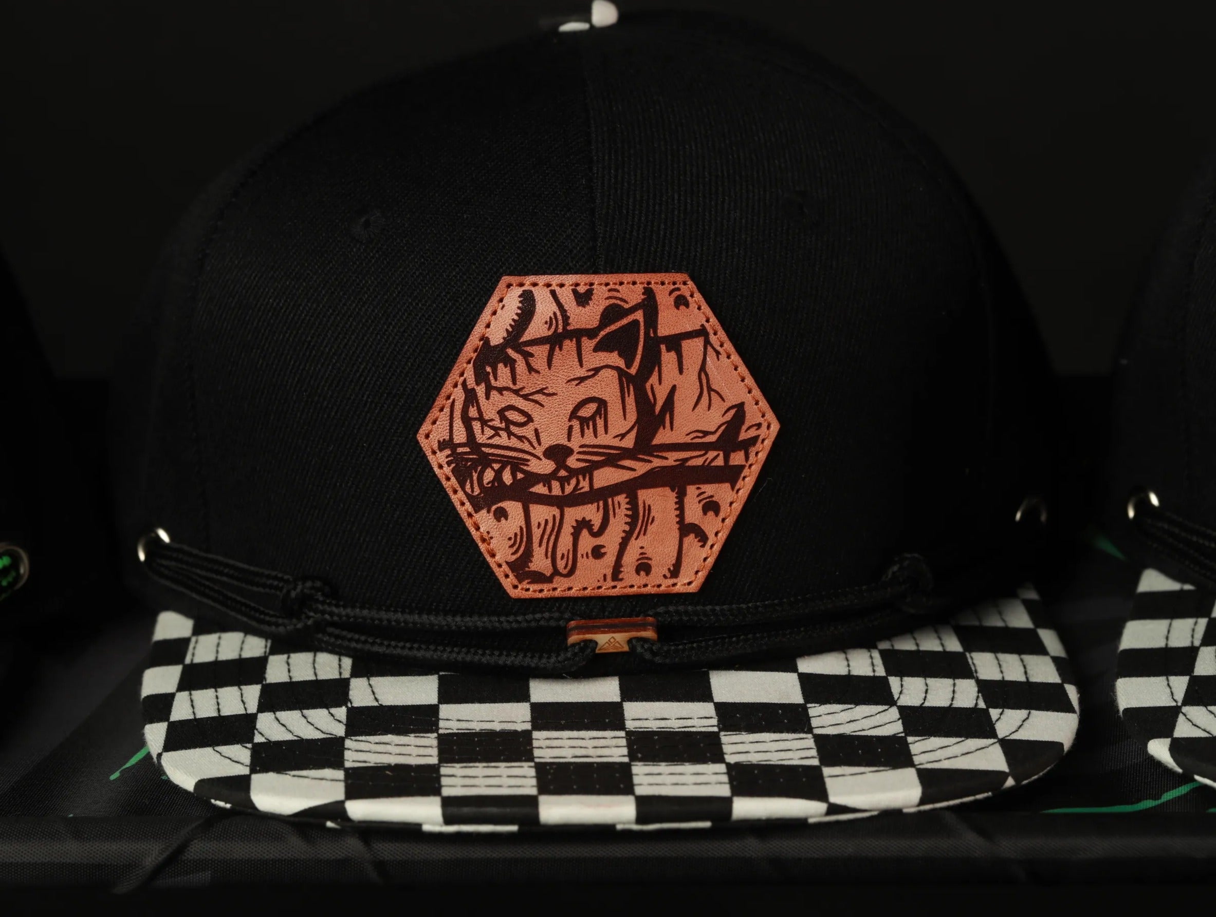 A close-up view of a black baseball cap with a checkered pattern and a leather patch featuring a cat design.