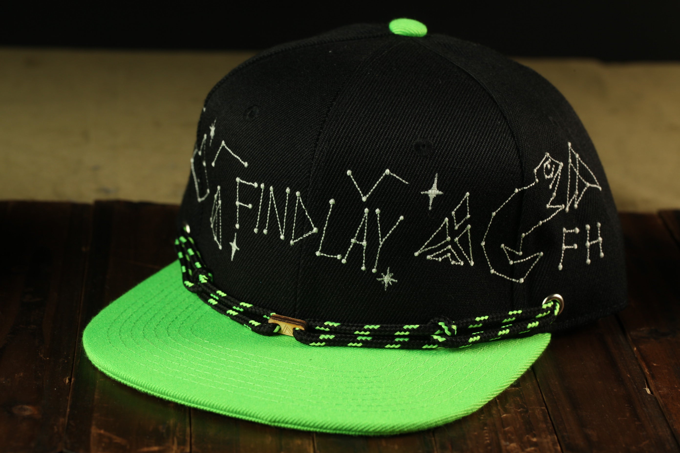 A black baseball cap with a green brim, featuring embroidered text and starry designs on the front.