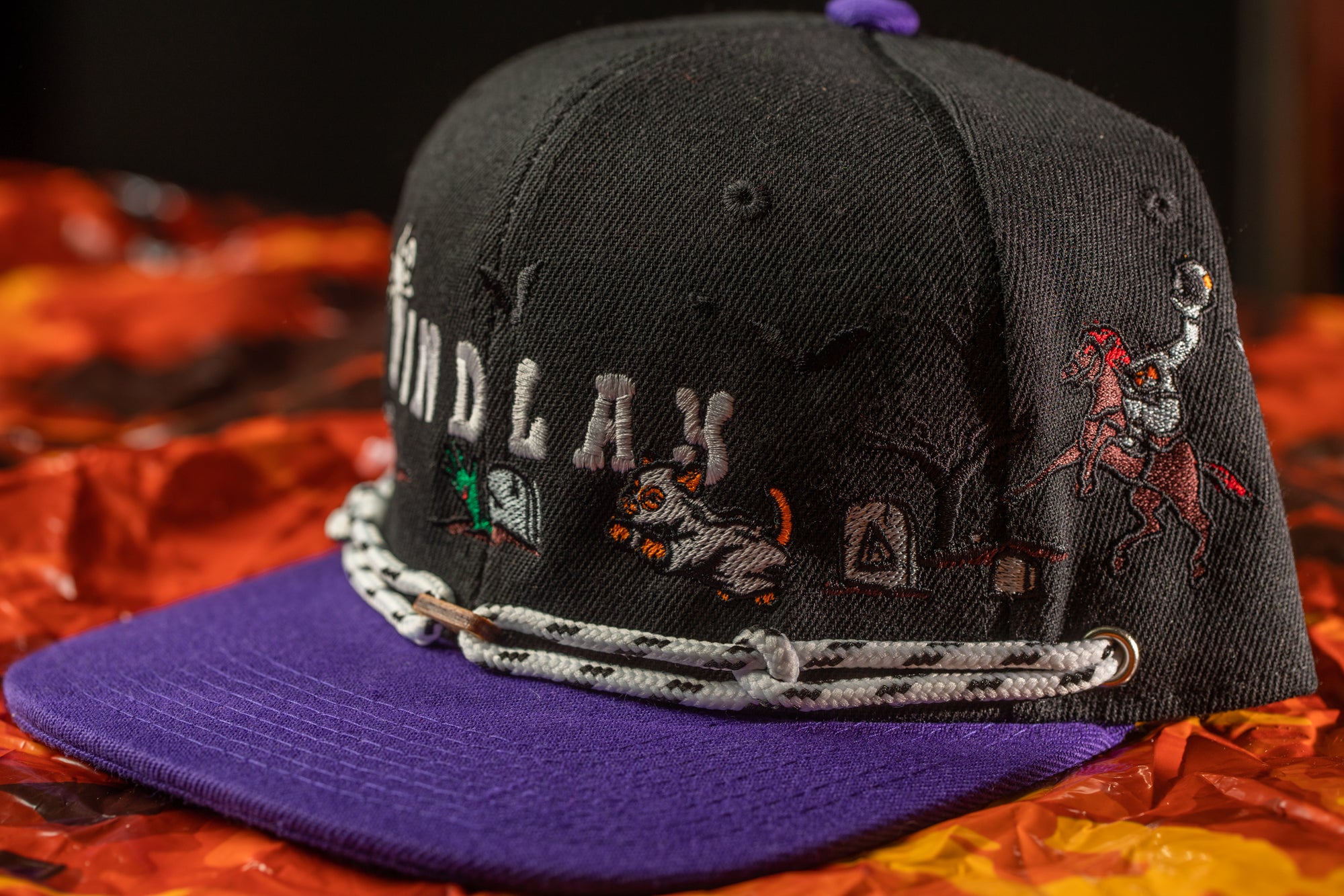 A black baseball cap with purple brim and various embroidered designs, including a character and a scene, is shown against a background of orange and red fabric.