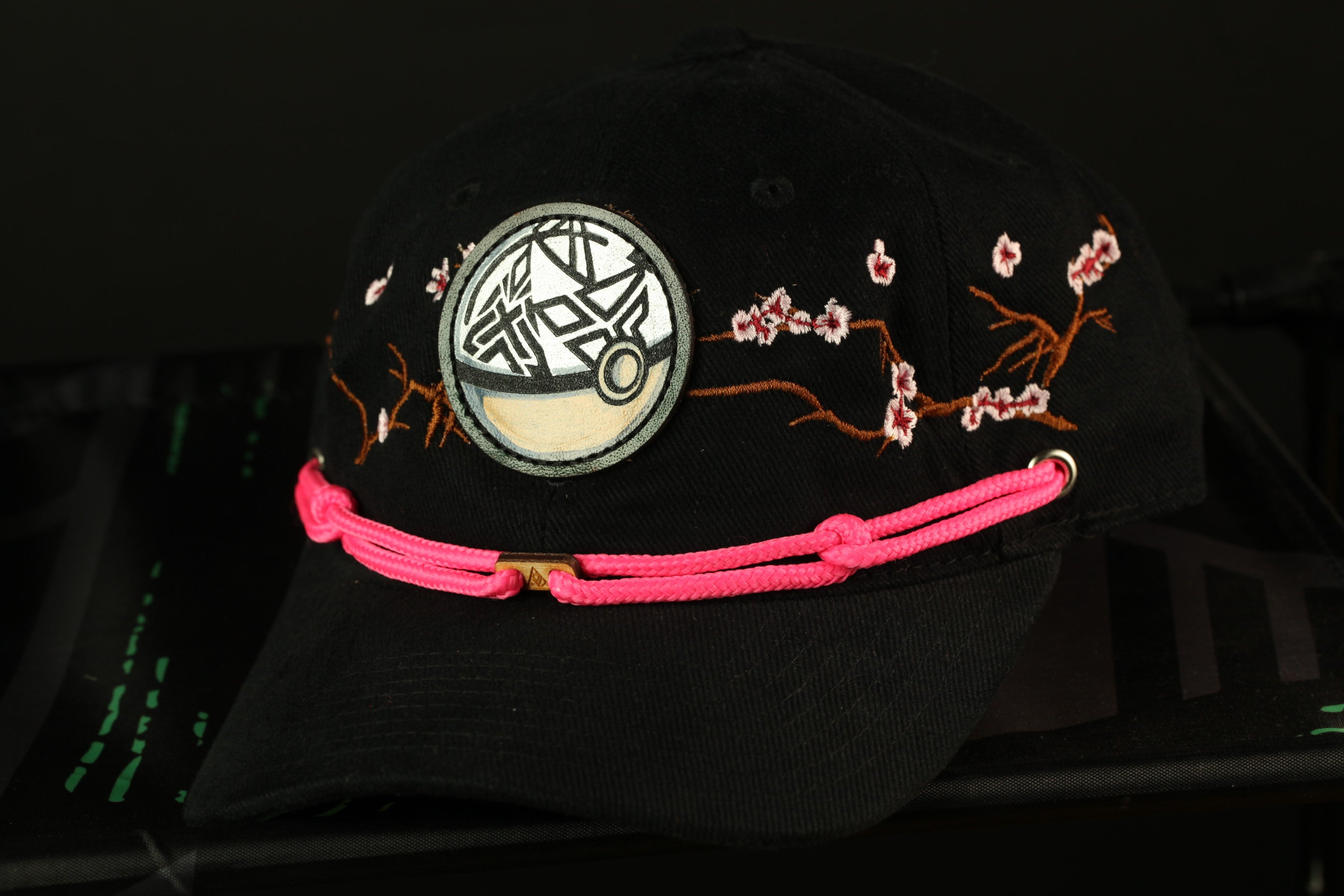A black baseball cap with a pink braided strap and a circular patch on the front, featuring a stylized design.