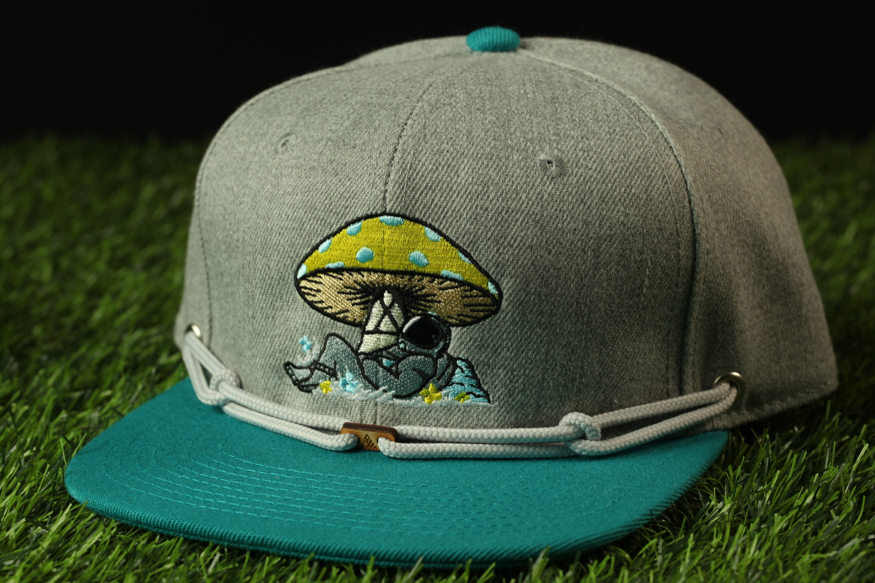 A gray baseball cap with a teal brim and a mushroom design on the front, placed on a grassy surface.