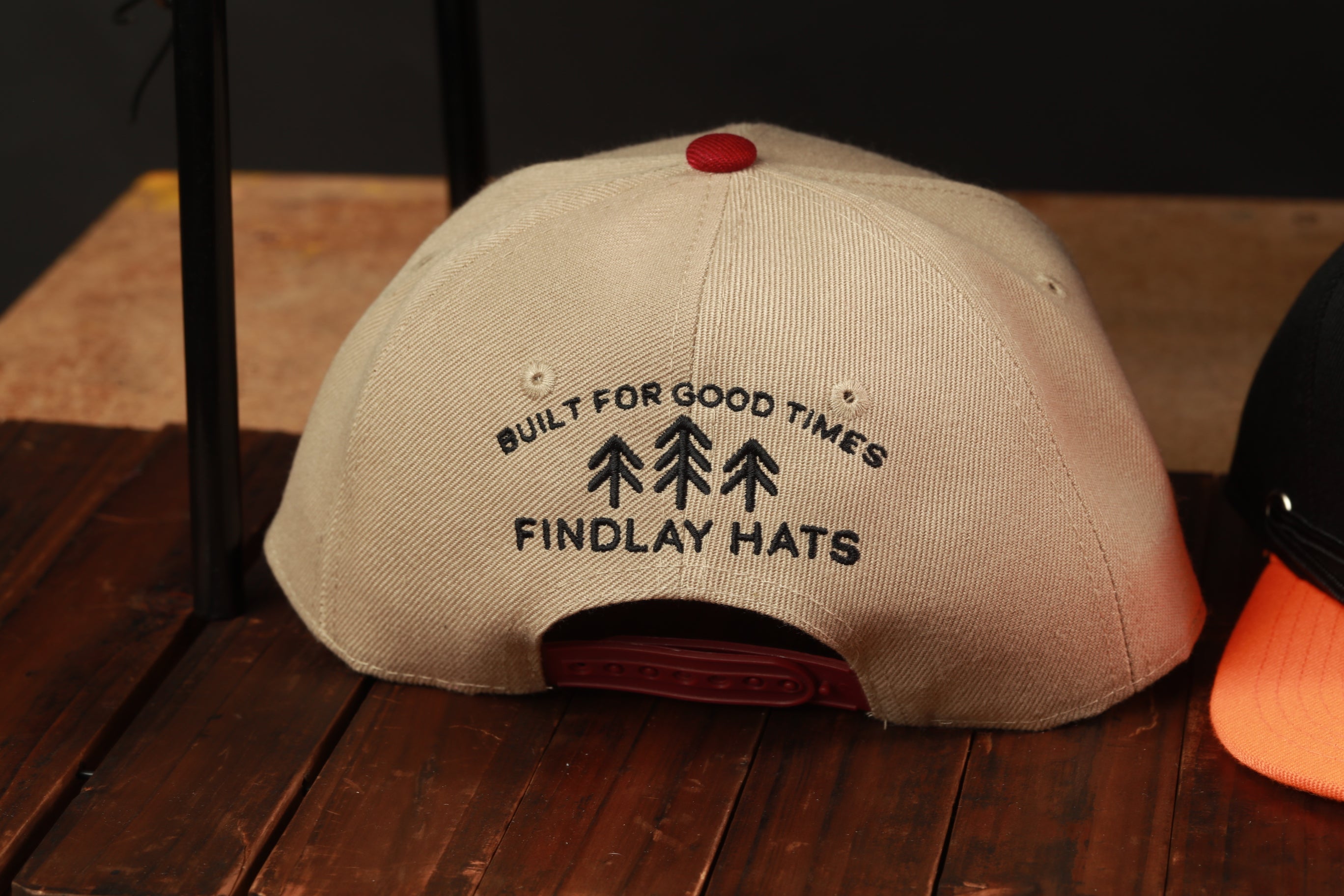 A beige baseball cap with the text "BUILT FOR GOOD TIMES FINDLAY HATS" printed on it, sitting on a wooden surface.