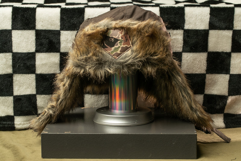 A fur-lined hat with a camouflage pattern is displayed on a stand against a black and white checkered background.