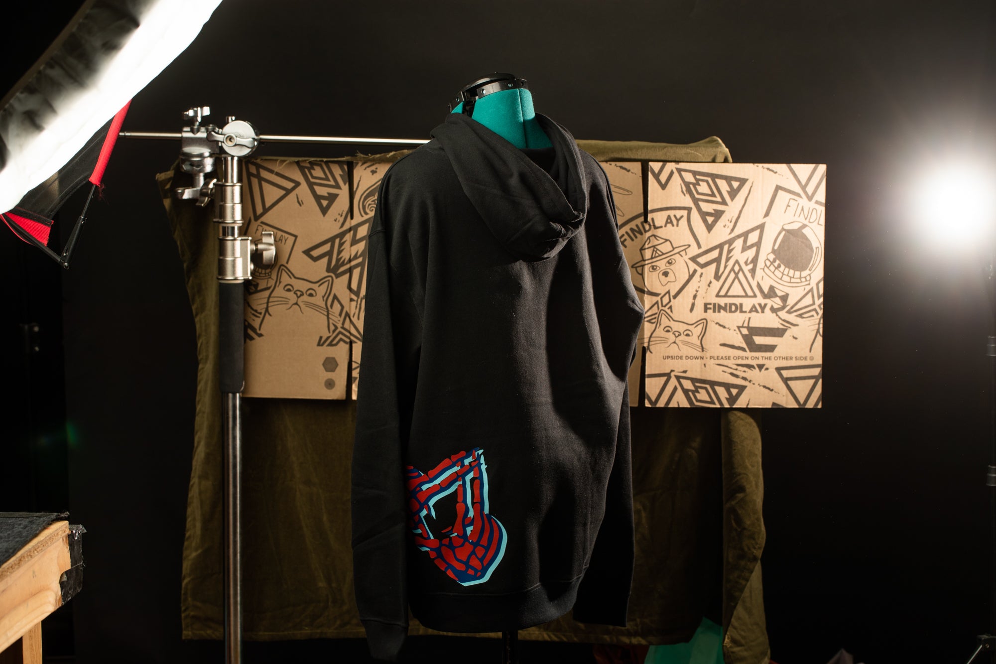 A black hoodie with a graphic design on the back, displayed on a mannequin in a dark room with a cardboard box and a light source in the background.
