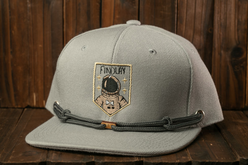 A gray baseball cap with a patch featuring an astronaut and the text "FINDLAY" on a wooden background.