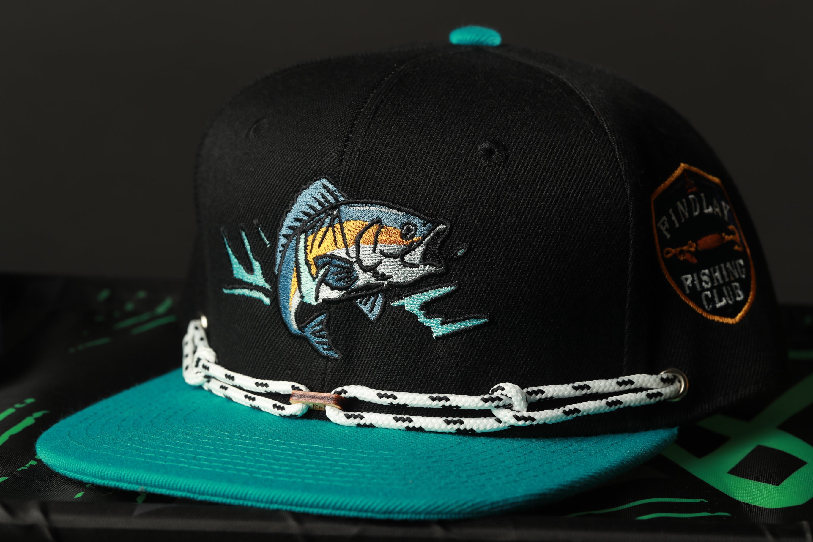 A black baseball cap with a teal brim and a fish embroidered on the front, sitting on a black surface.