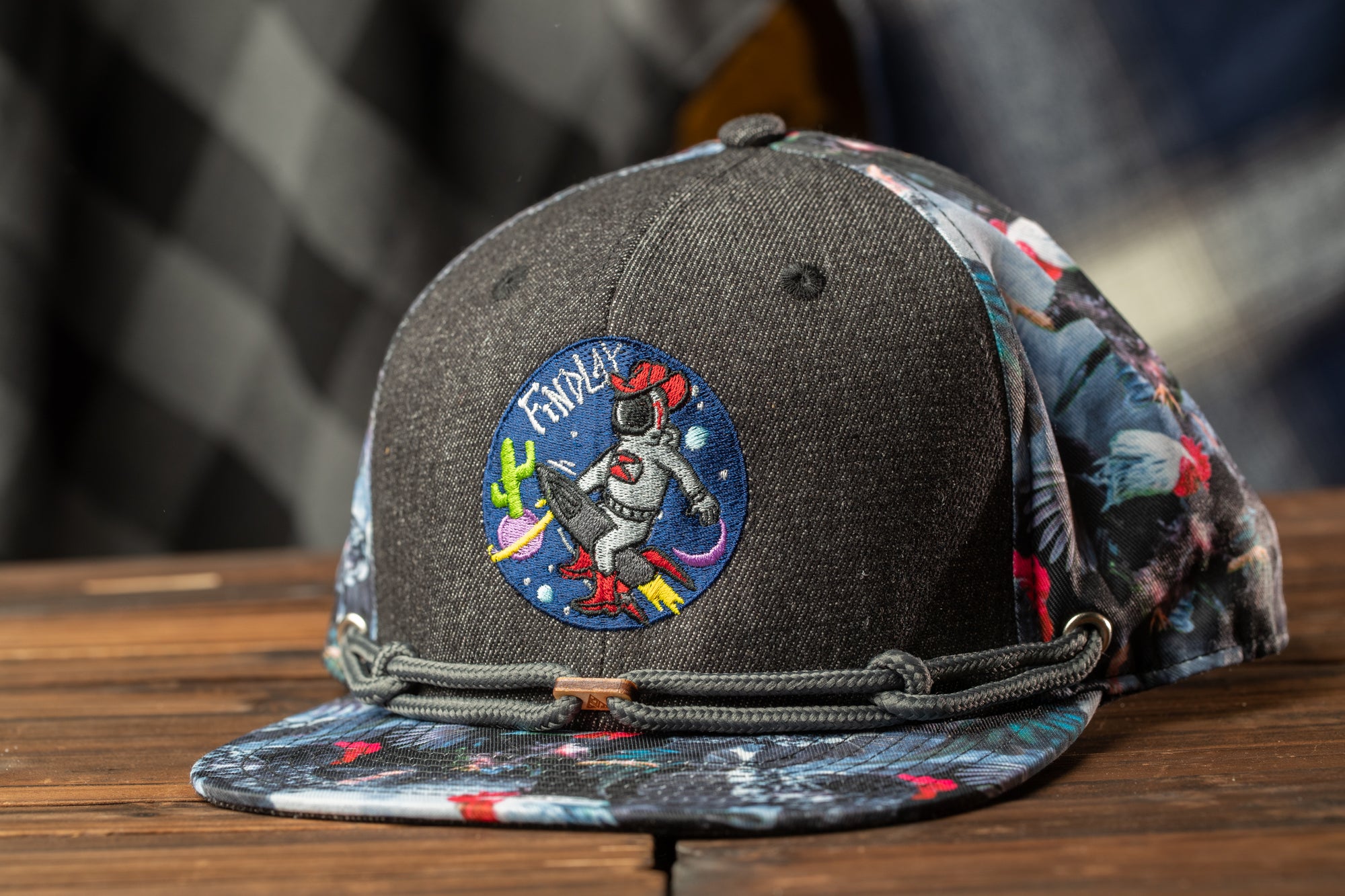 A black baseball cap with a colorful, embroidered design on the front, featuring a space-themed motif with a cactus and a rocket ship.