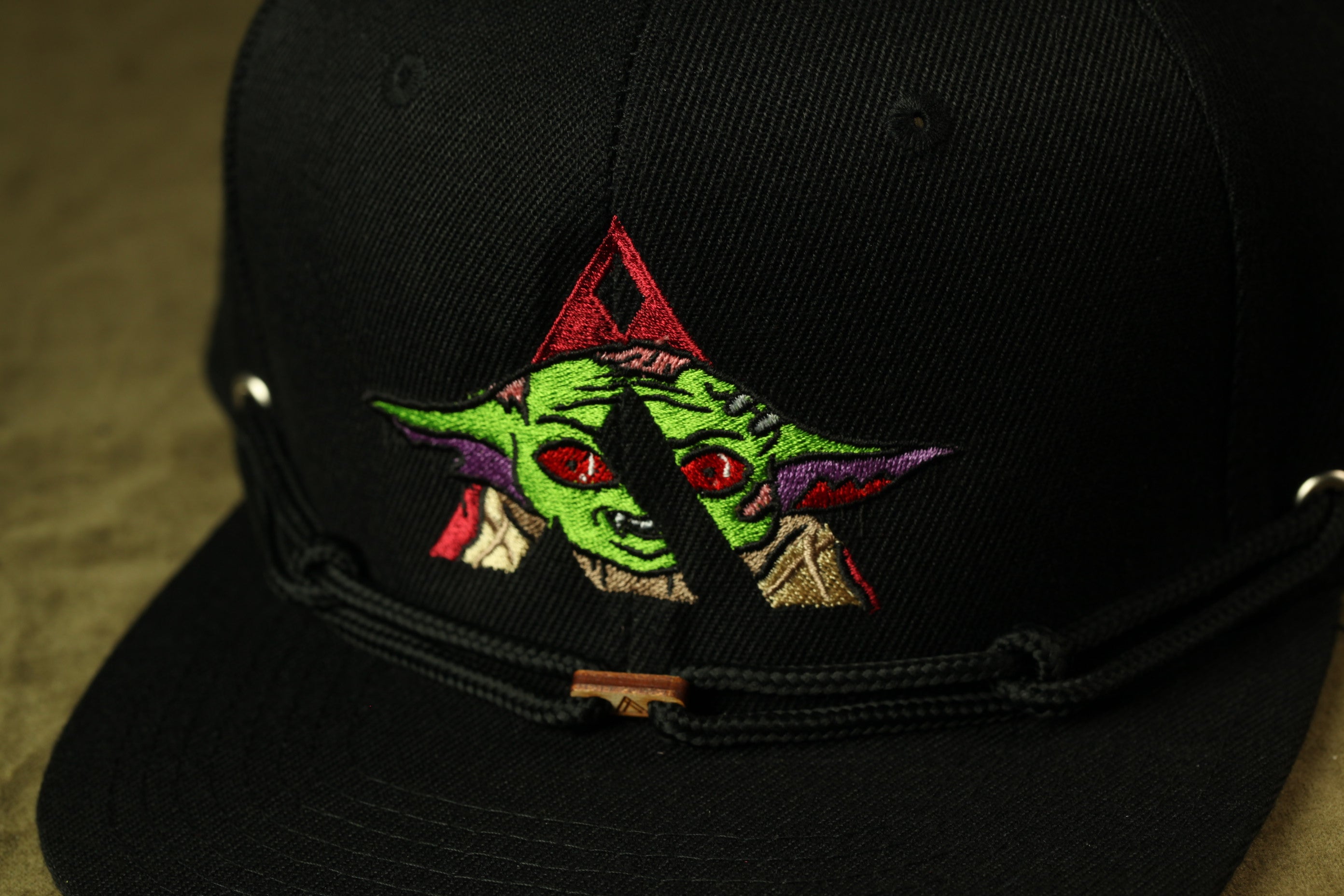 A close-up view of a black baseball cap with a colorful embroidered design featuring a stylized character resembling a green alien or alien-like creature.