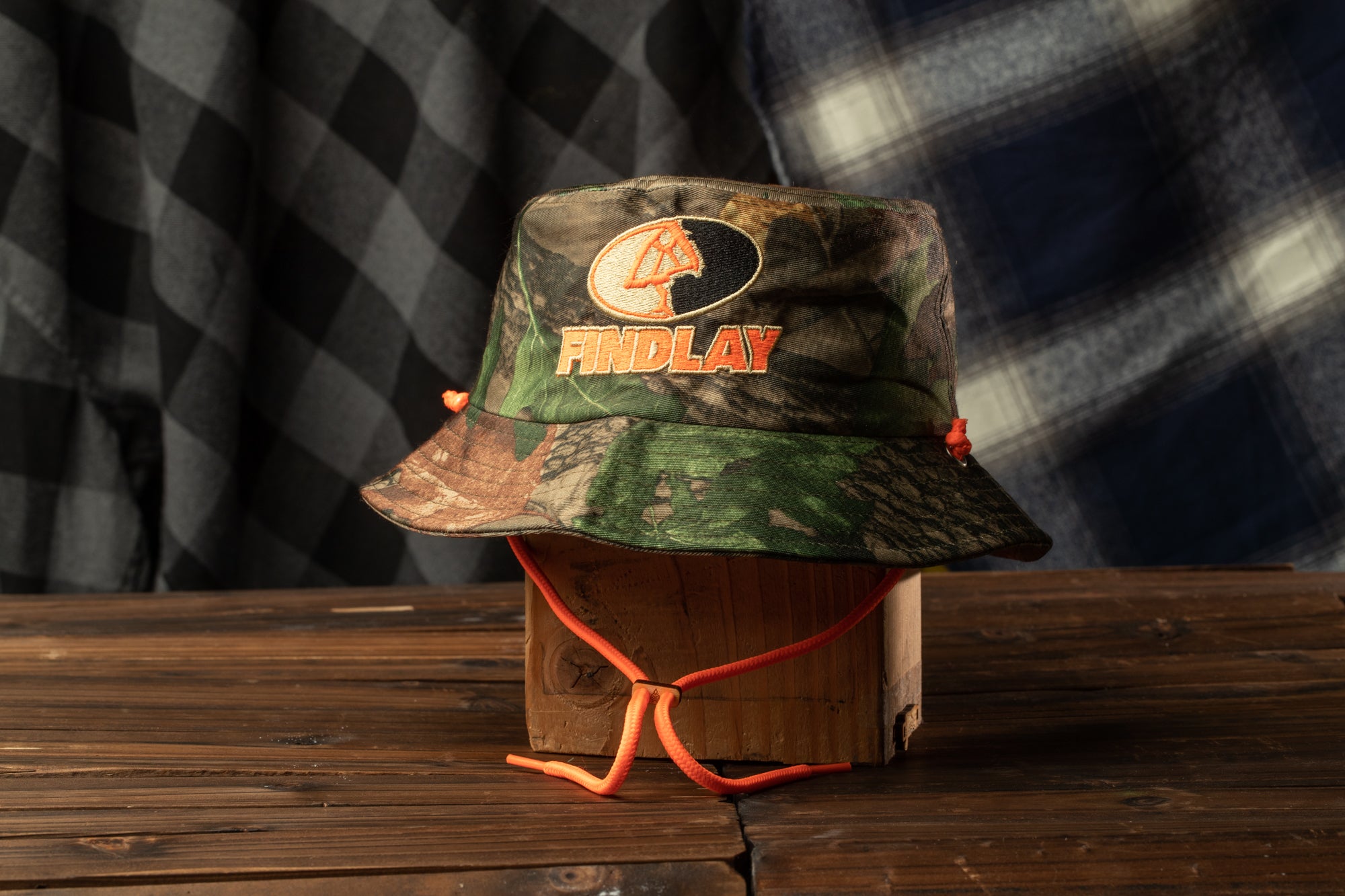A camouflage bucket hat with the word "FIND LAY" printed on it, sitting on a wooden surface.