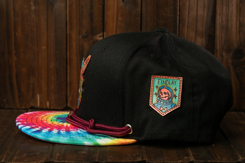 A black baseball cap with a colorful tie-dye pattern and a patch on the front, placed on a wooden surface.