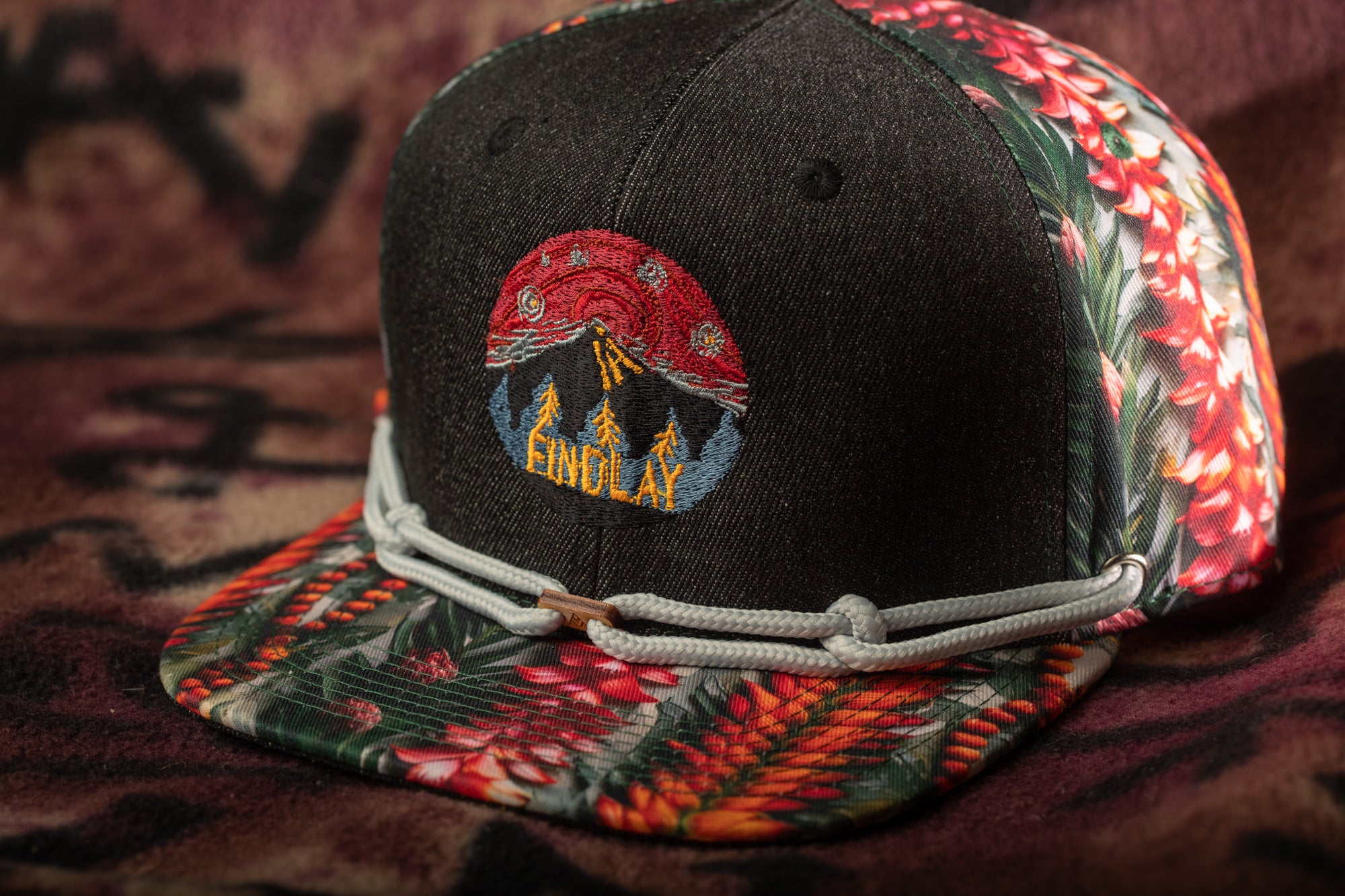 A black baseball cap with a colorful floral pattern and a red and yellow logo on the front.