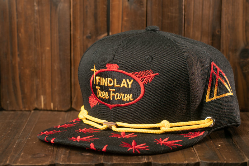 A black baseball cap with red and yellow embroidered designs, including a tree farm logo, is displayed on a wooden surface.