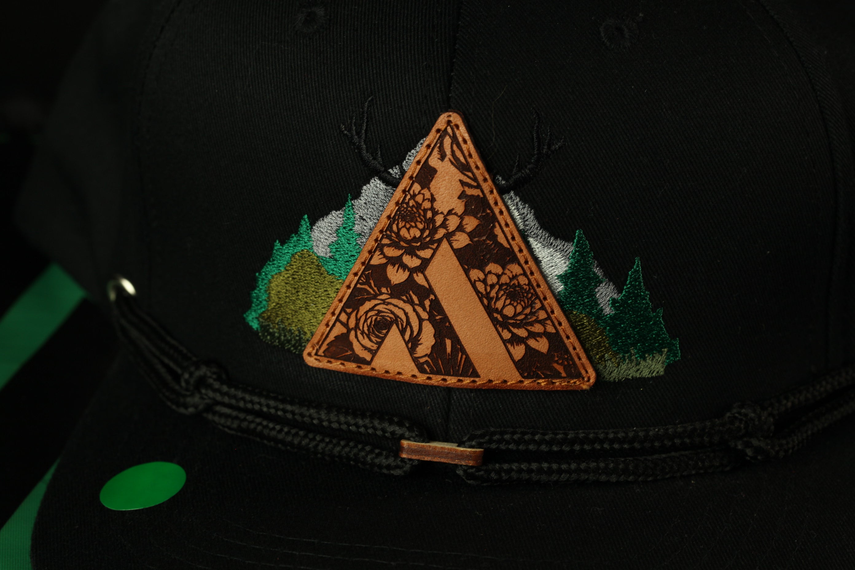 A close-up view of a black baseball cap with a decorative triangular patch featuring a floral design and a mountain landscape.