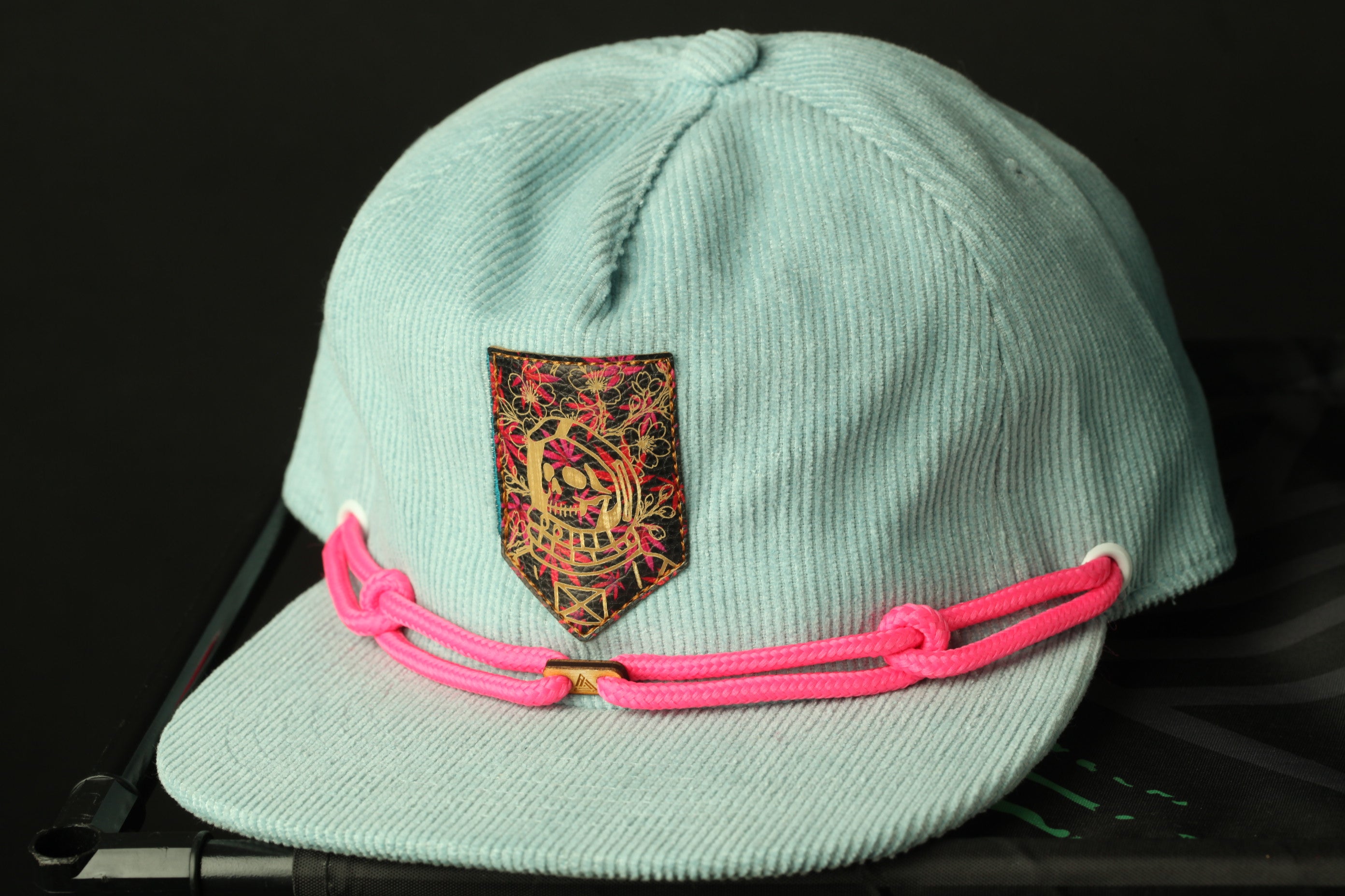 A light blue baseball cap with a decorative patch and pink braided straps is shown in the image.