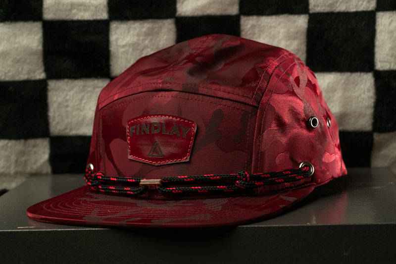 A red camouflage baseball cap with the word "INDLAY" printed on it, sitting on a black surface against a black and white checkered background.