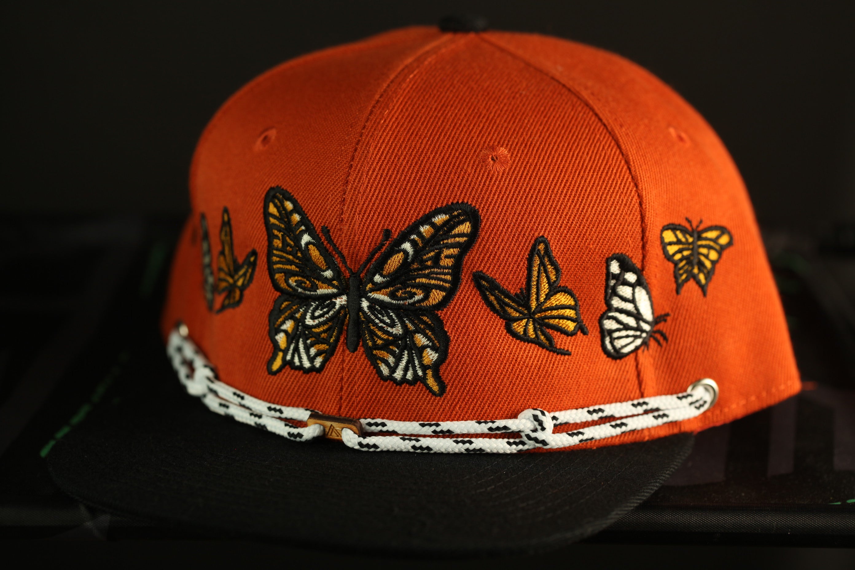 An orange baseball cap with a black brim and white braided strap, decorated with a pattern of butterflies.
