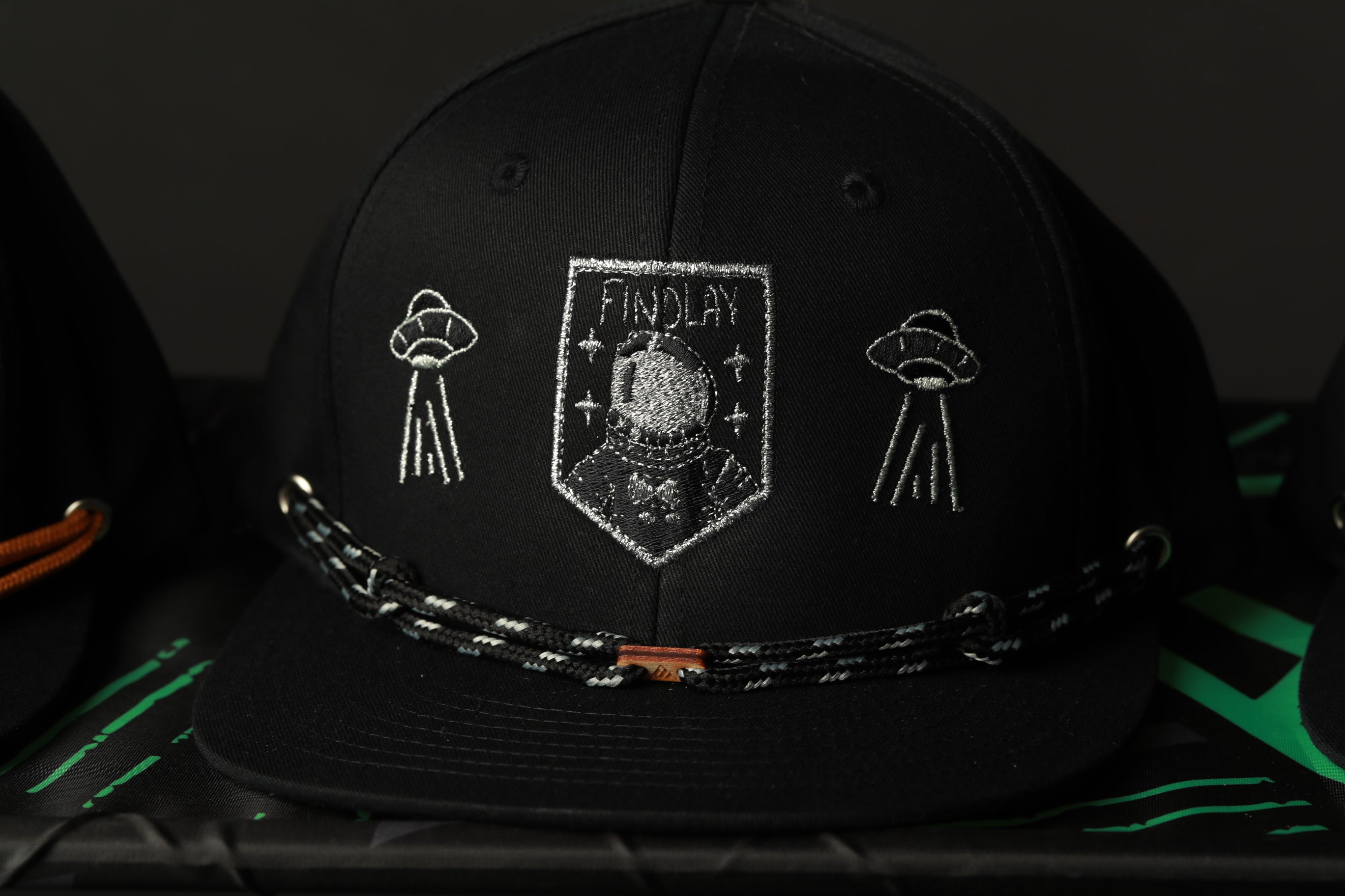 A black baseball cap with a white embroidered design on the front, featuring a shield-shaped emblem with a figure and two alien-like figures on either side.