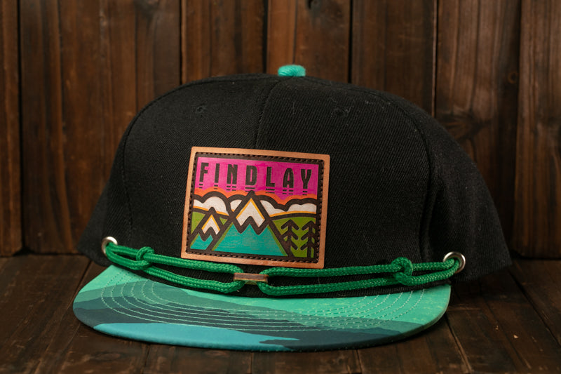 A black baseball cap with a colorful graphic patch on the front, featuring a mountain landscape design.