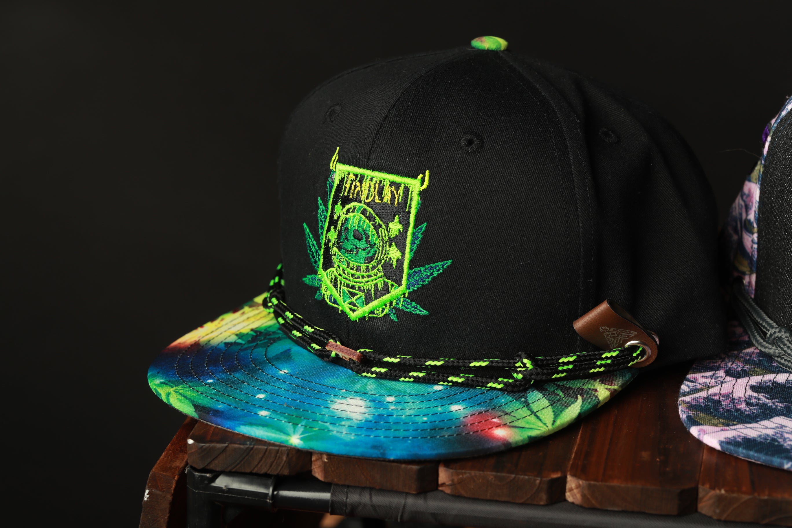 A black baseball cap with a colorful tie-dye pattern and a green and yellow embroidered logo on the front.