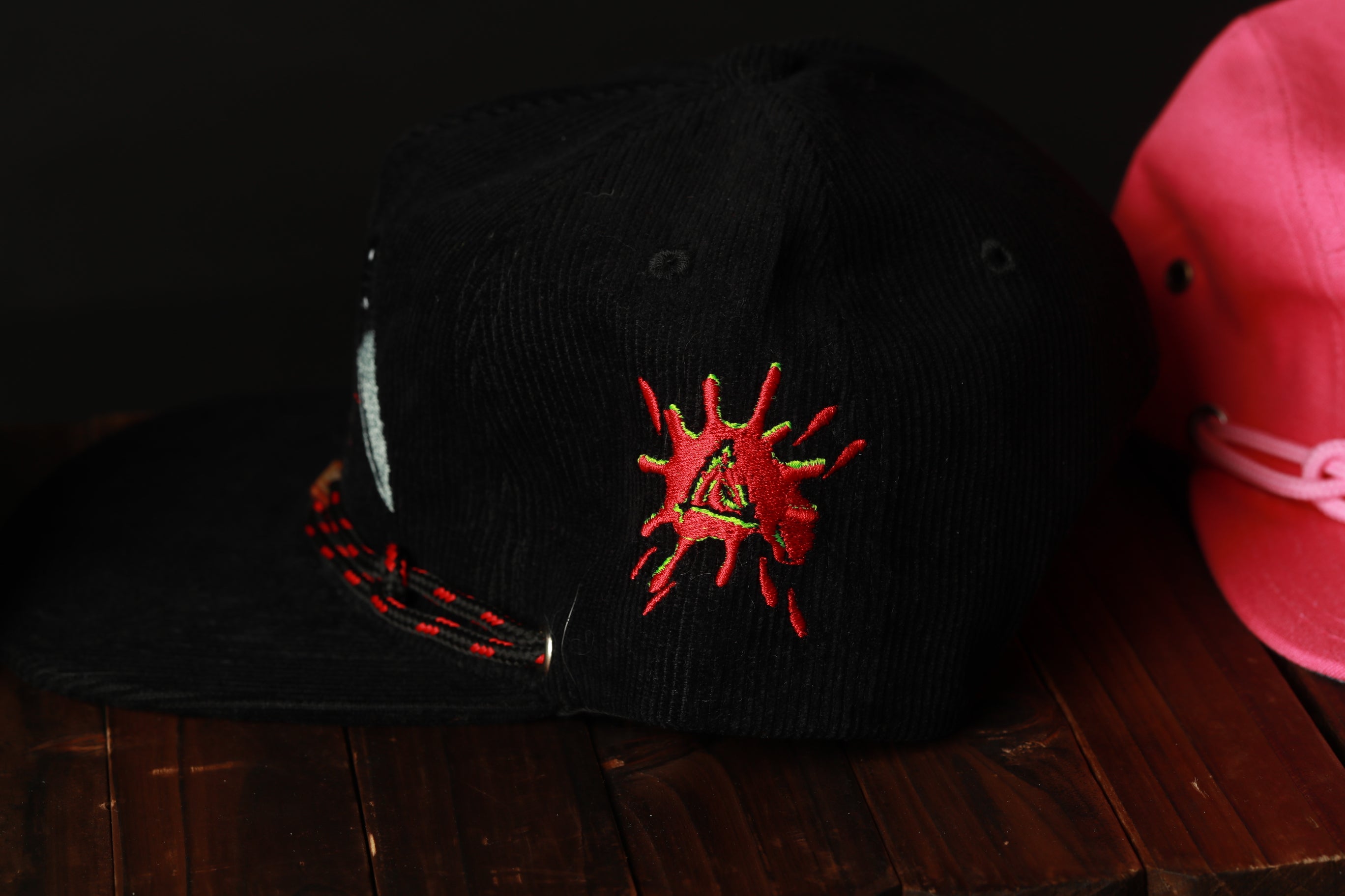 a black baseball cap with a red and yellow embroidered design on the front, placed on a wooden surface next to a pink baseball cap.