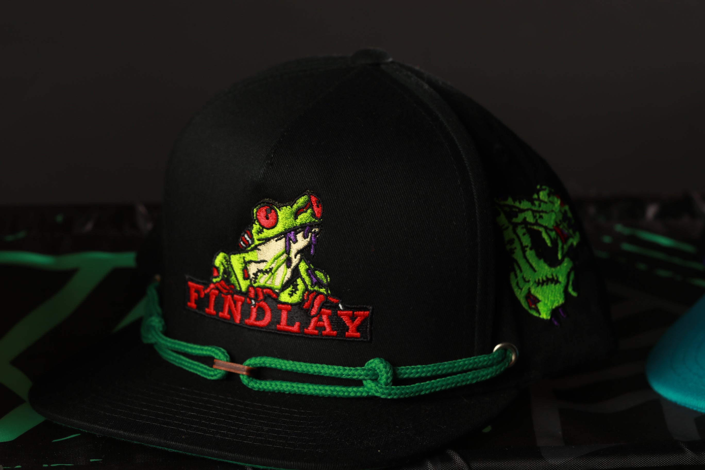 A black baseball cap with a green and red frog design and the word "FIND LAY" printed on it.