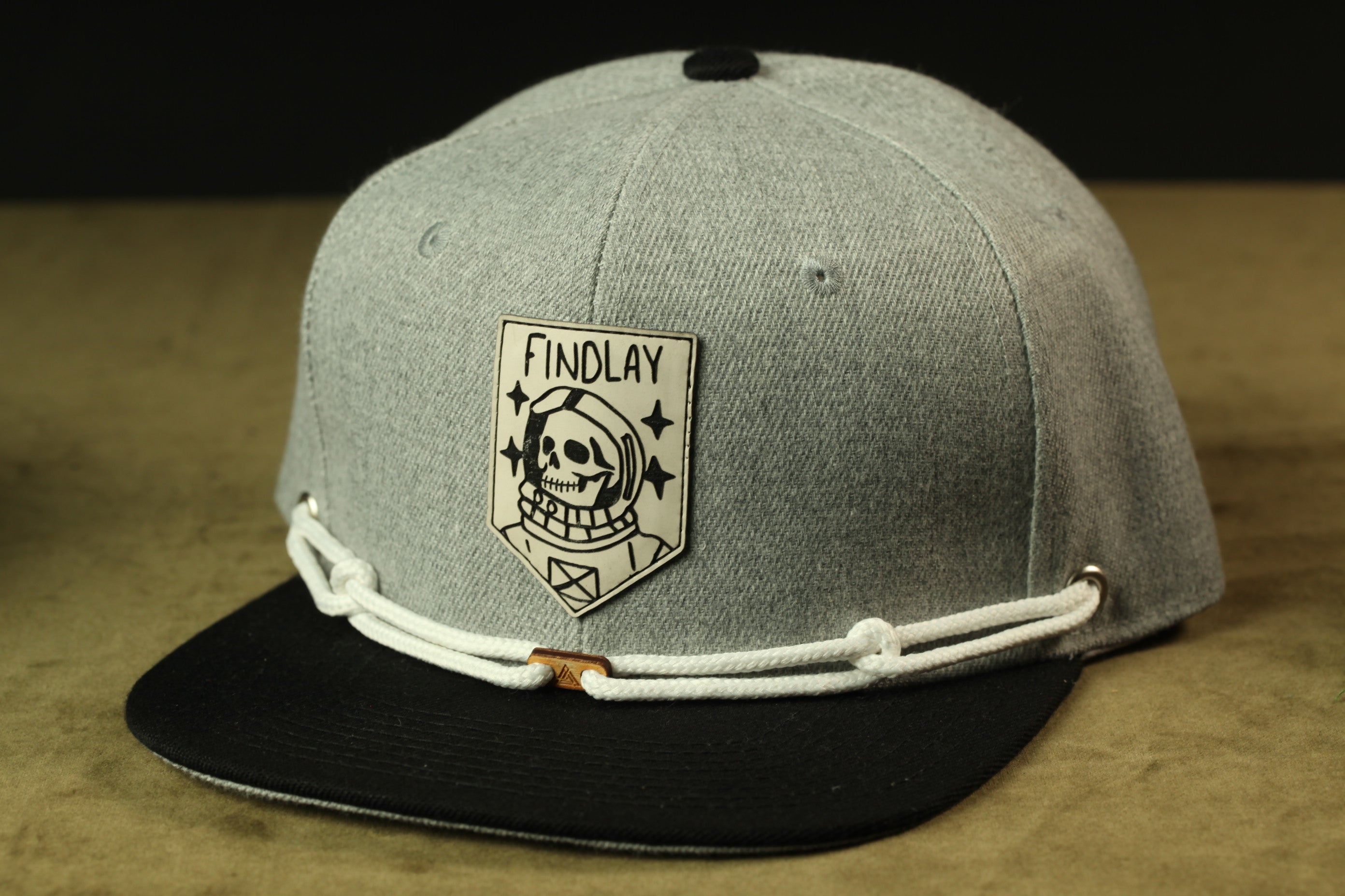 A gray baseball cap with a black brim and a white band around the front. The cap has a patch on the front that says "FINDLAY" and features a skull and crossbones design.