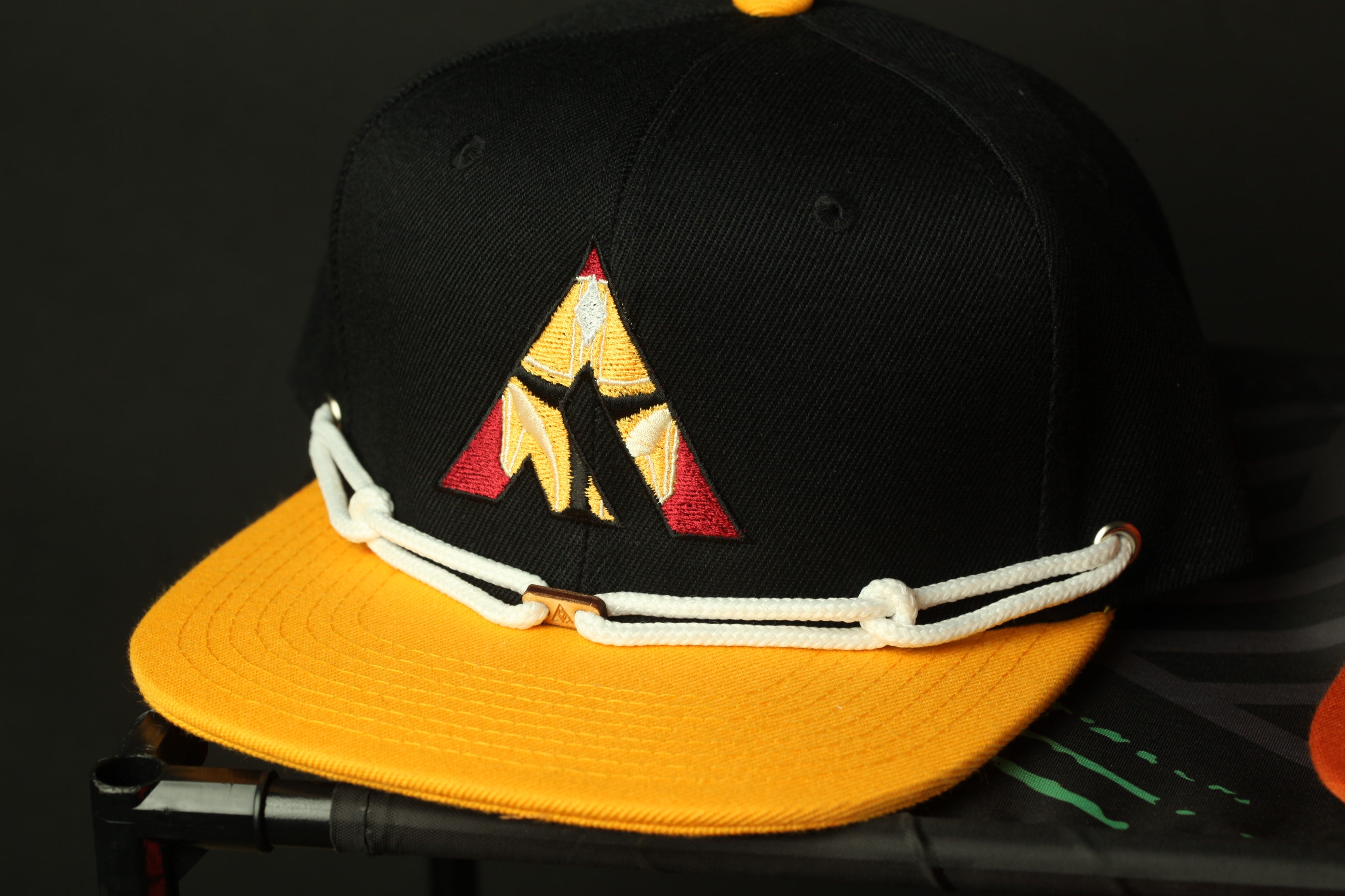 A black baseball cap with a yellow brim and a red and yellow logo on the front, sitting on a black surface.