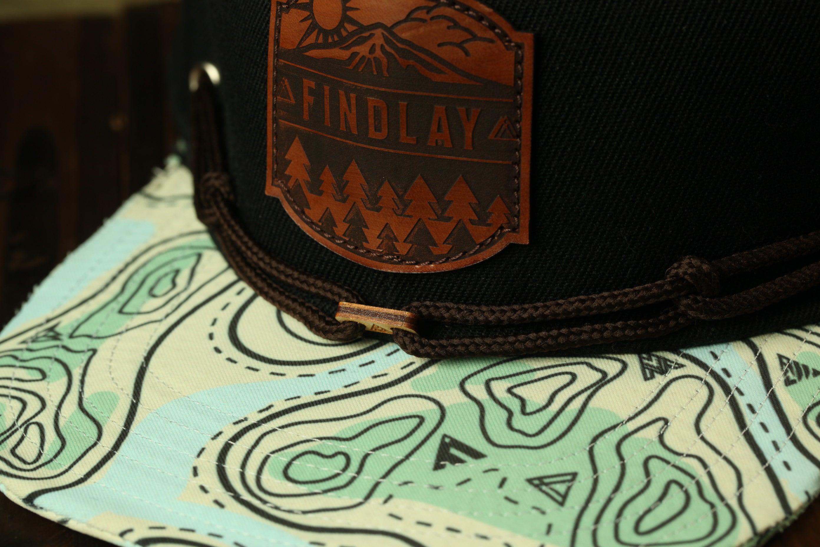 A close-up view of a black baseball cap with a brown leather patch on the front, featuring a mountain landscape design.
