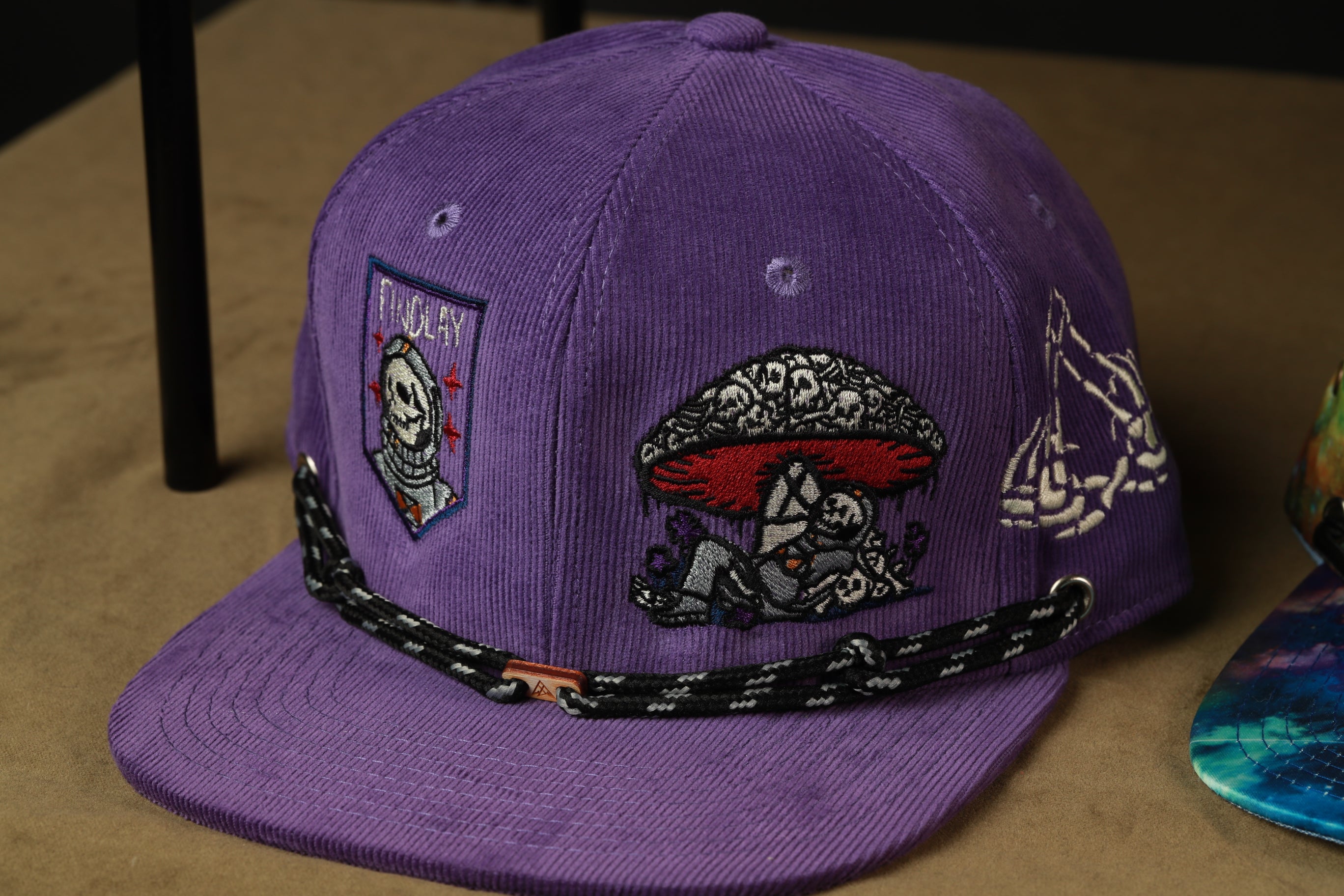A purple baseball cap with various embroidered designs, including a skull and crossbones, mushrooms, and other abstract patterns.