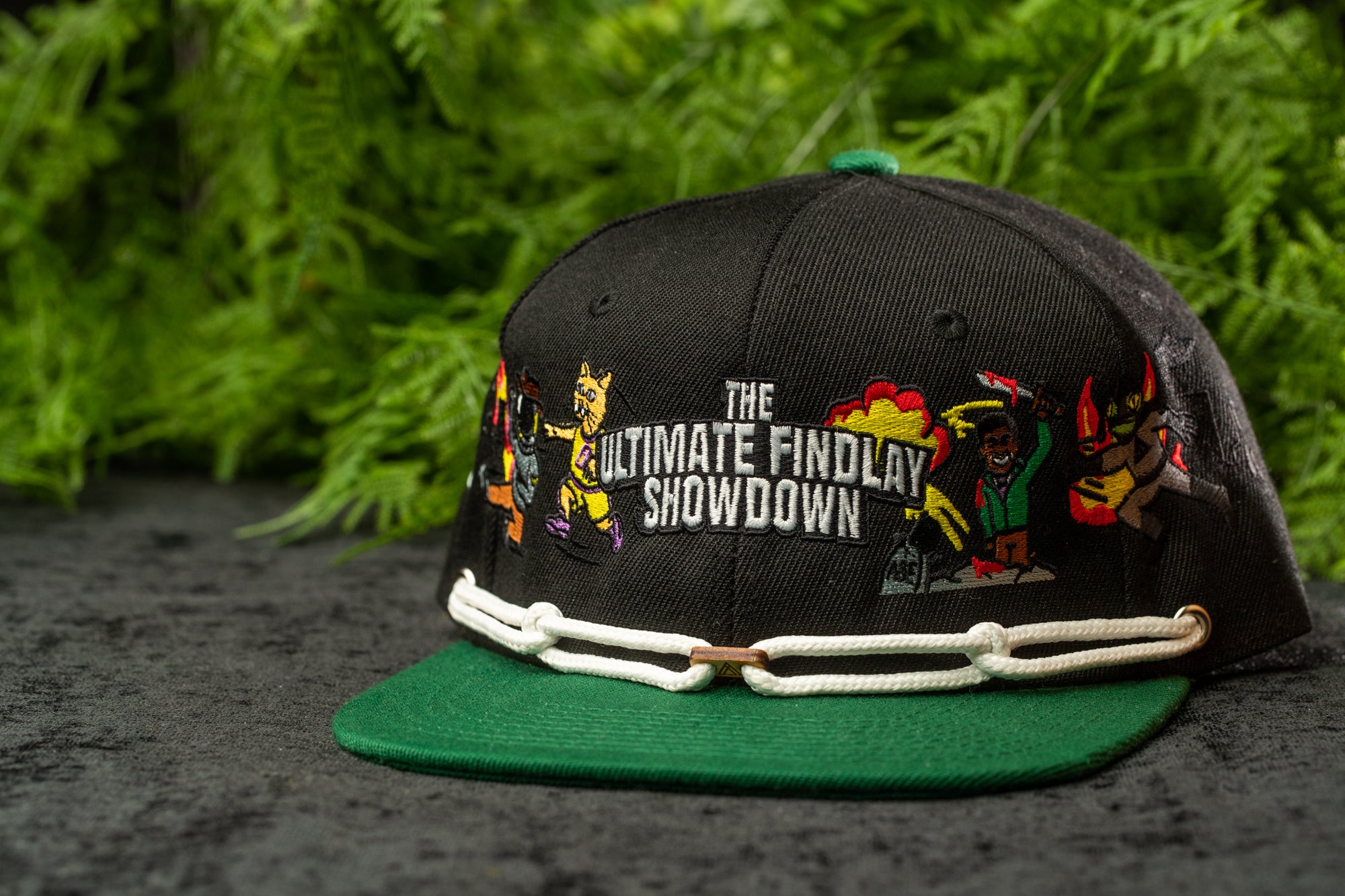 A black baseball cap with colorful cartoon characters and text on it, placed on a green brim.