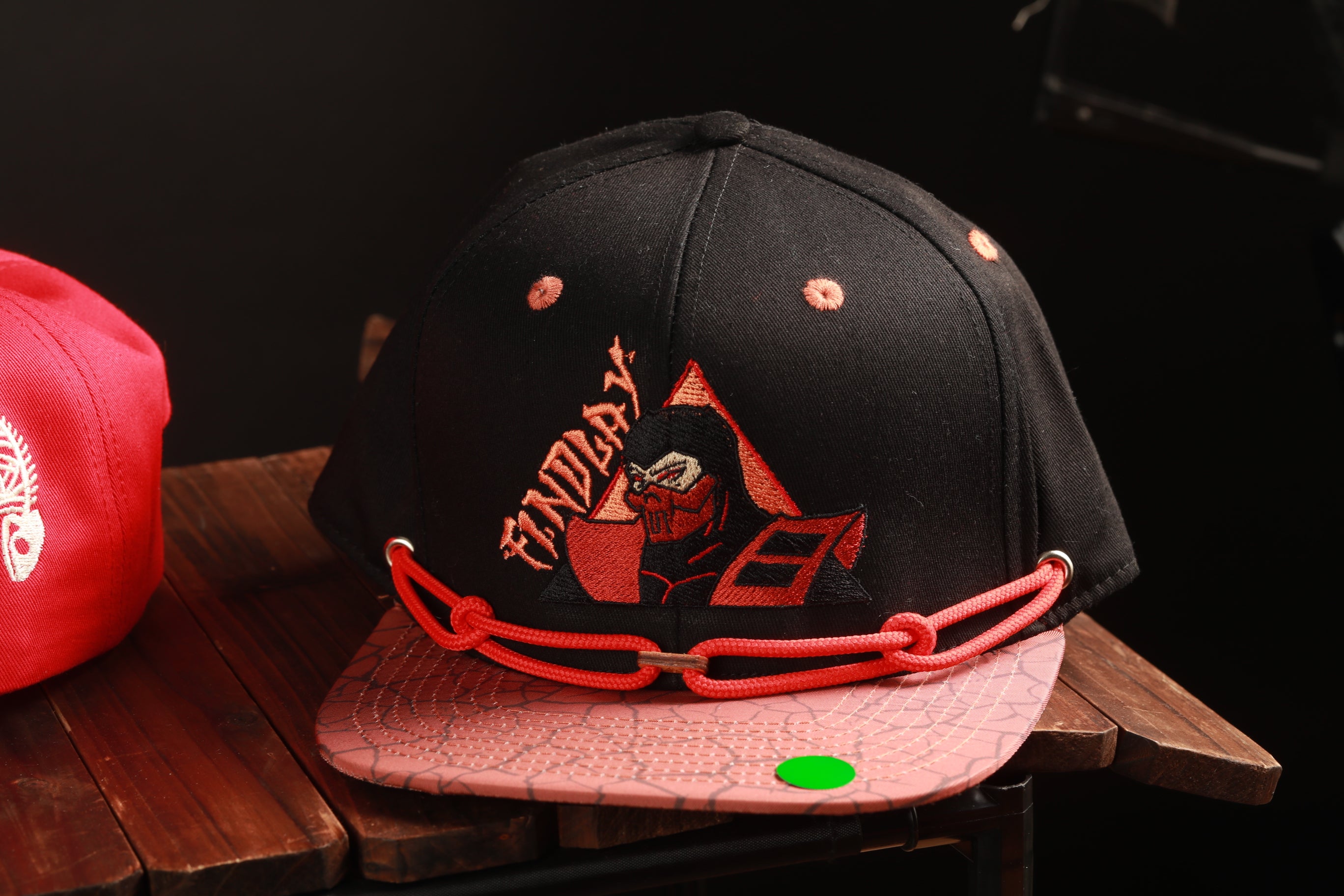 A black baseball cap with a pink brim and a red chain strap, featuring an embroidered design of a character, is displayed on a wooden surface.