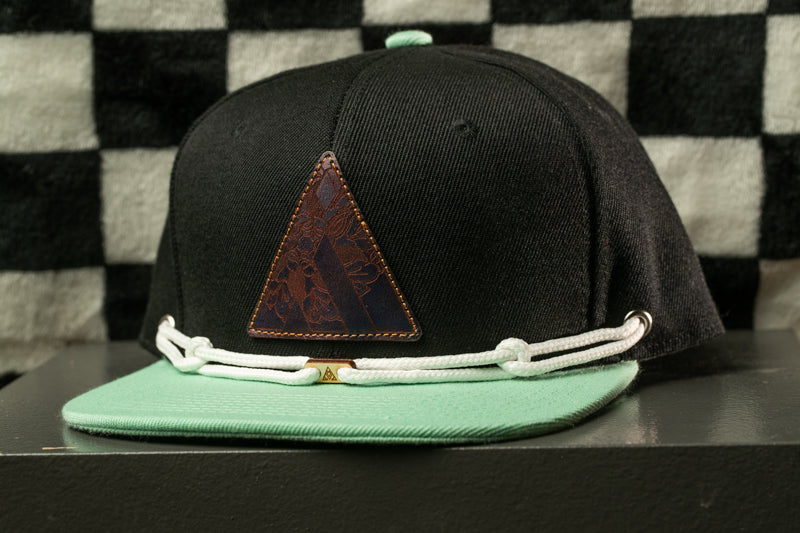A black baseball cap with a green brim and a brown leather patch on the front, displayed on a black surface against a checkered background.